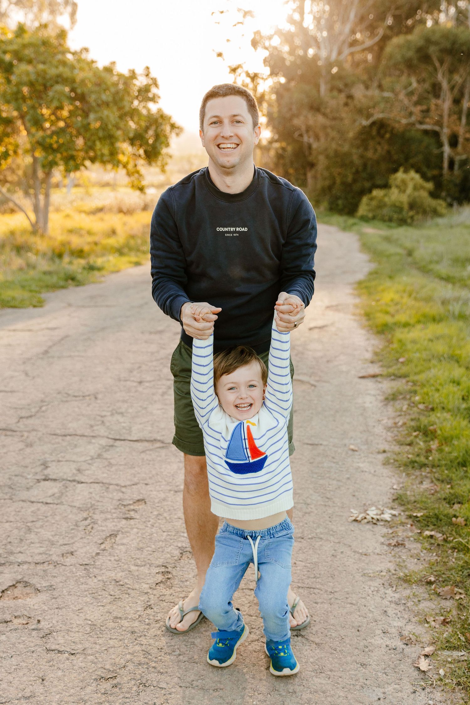 A smiling father holds his laughing young son's hands on a sunny outdoor dirt path.