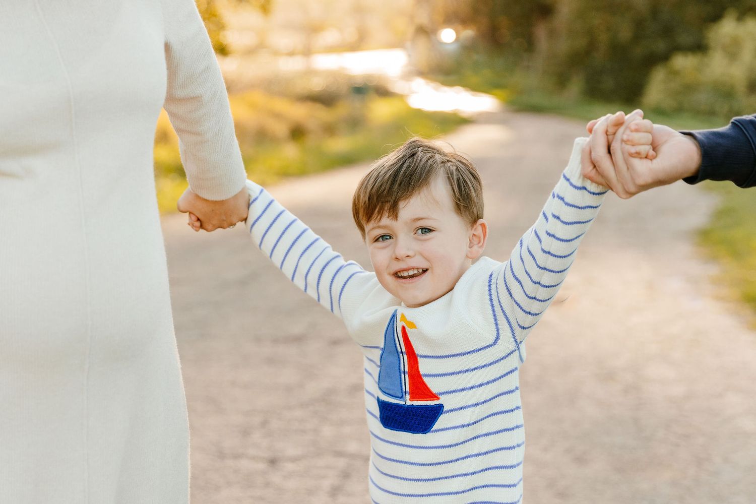 Excited young boy in striped sailboat shirt being swung by parents hands on a sunny path.
