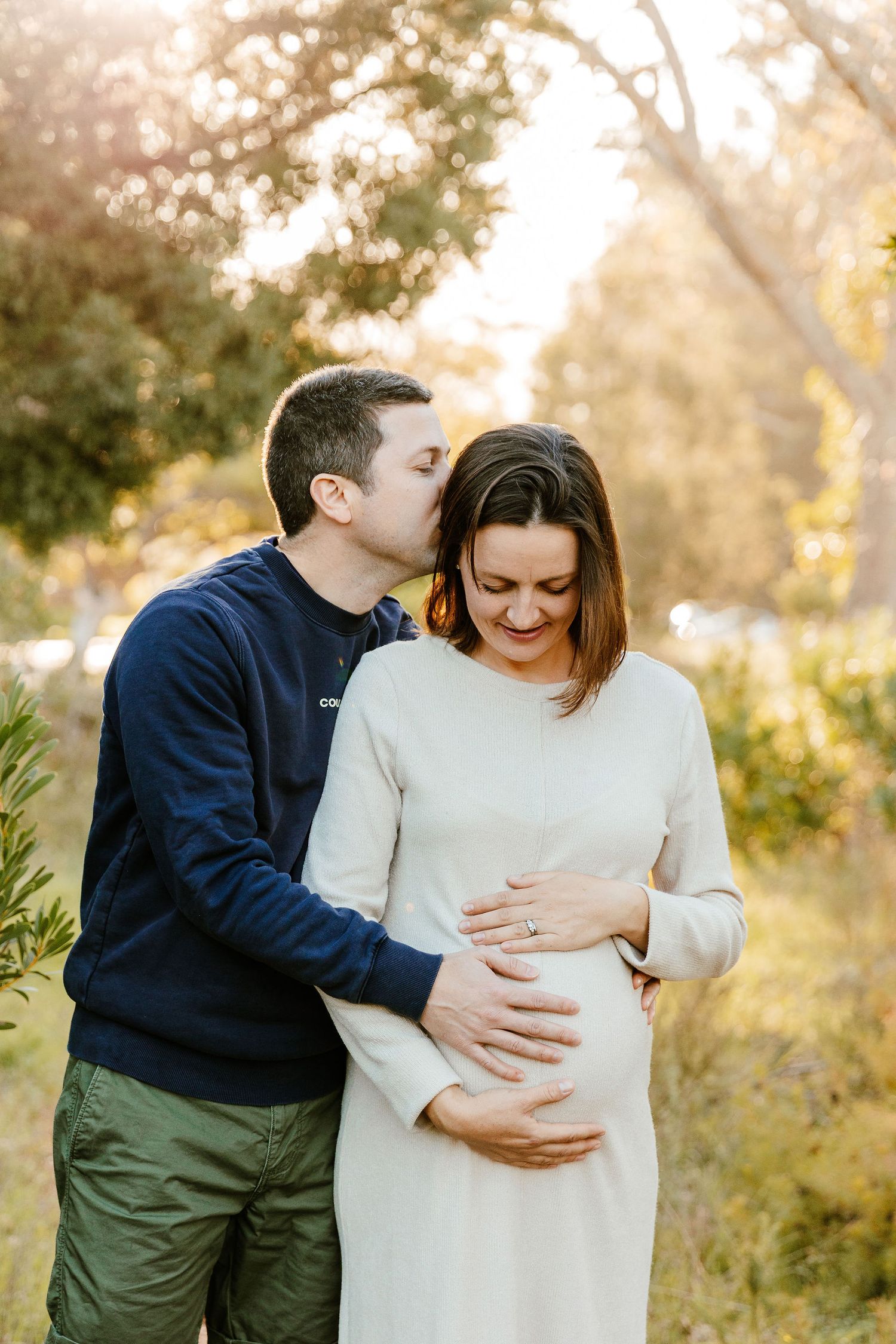 A man kisses a pregnant woman on the cheek while holding her baby bump outdoors in golden sunlight.