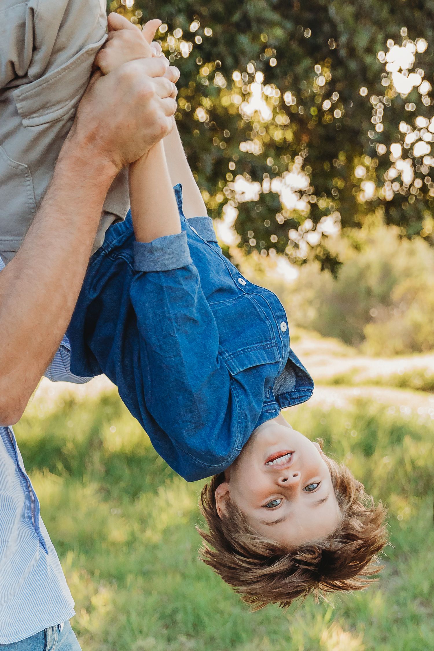 A joyful child in a denim shirt being held upside down by an adult outdoors in a sunny green park.