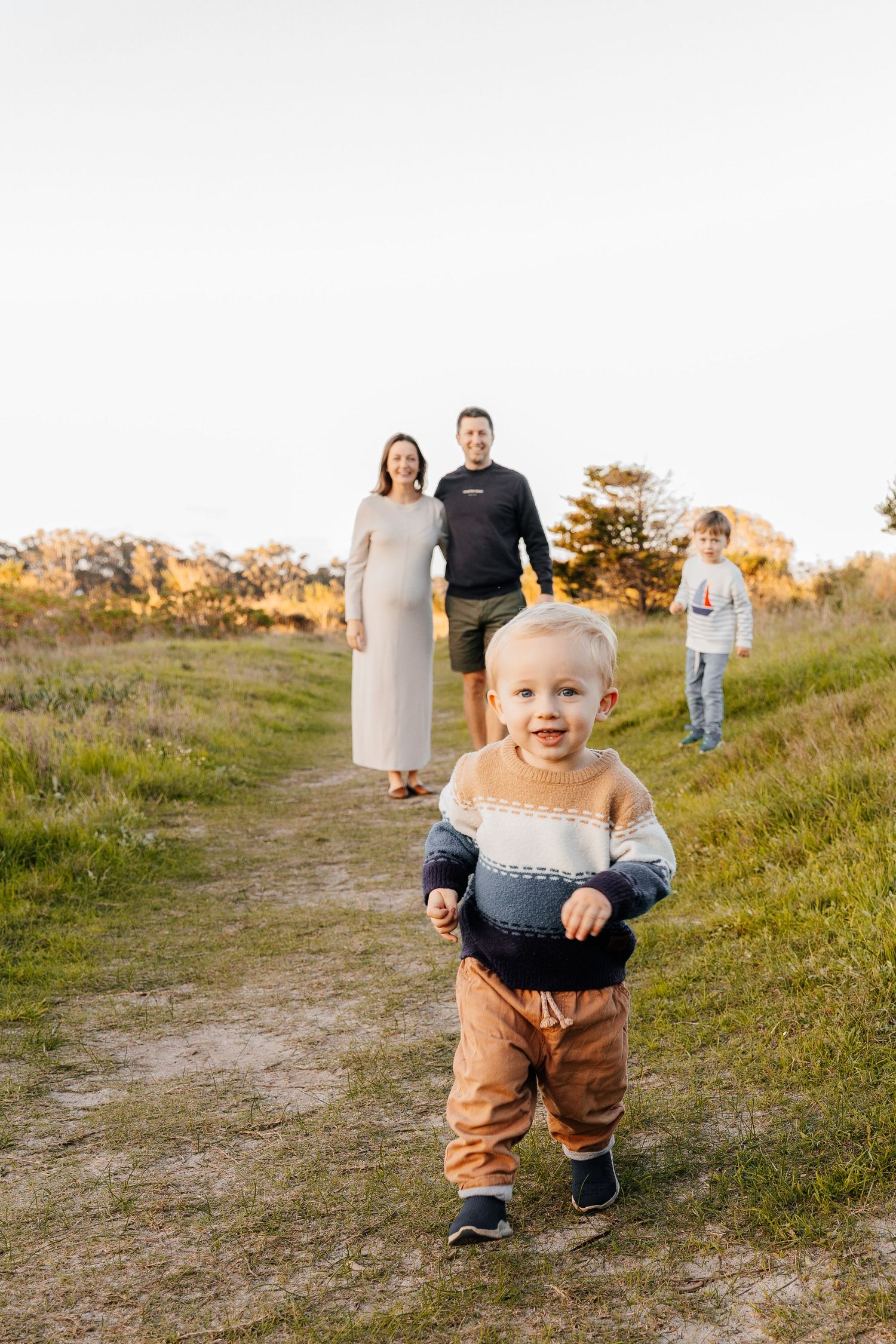 A toddler runs toward the camera on a grassy path as a pregnant woman, man, and child stand behind him.