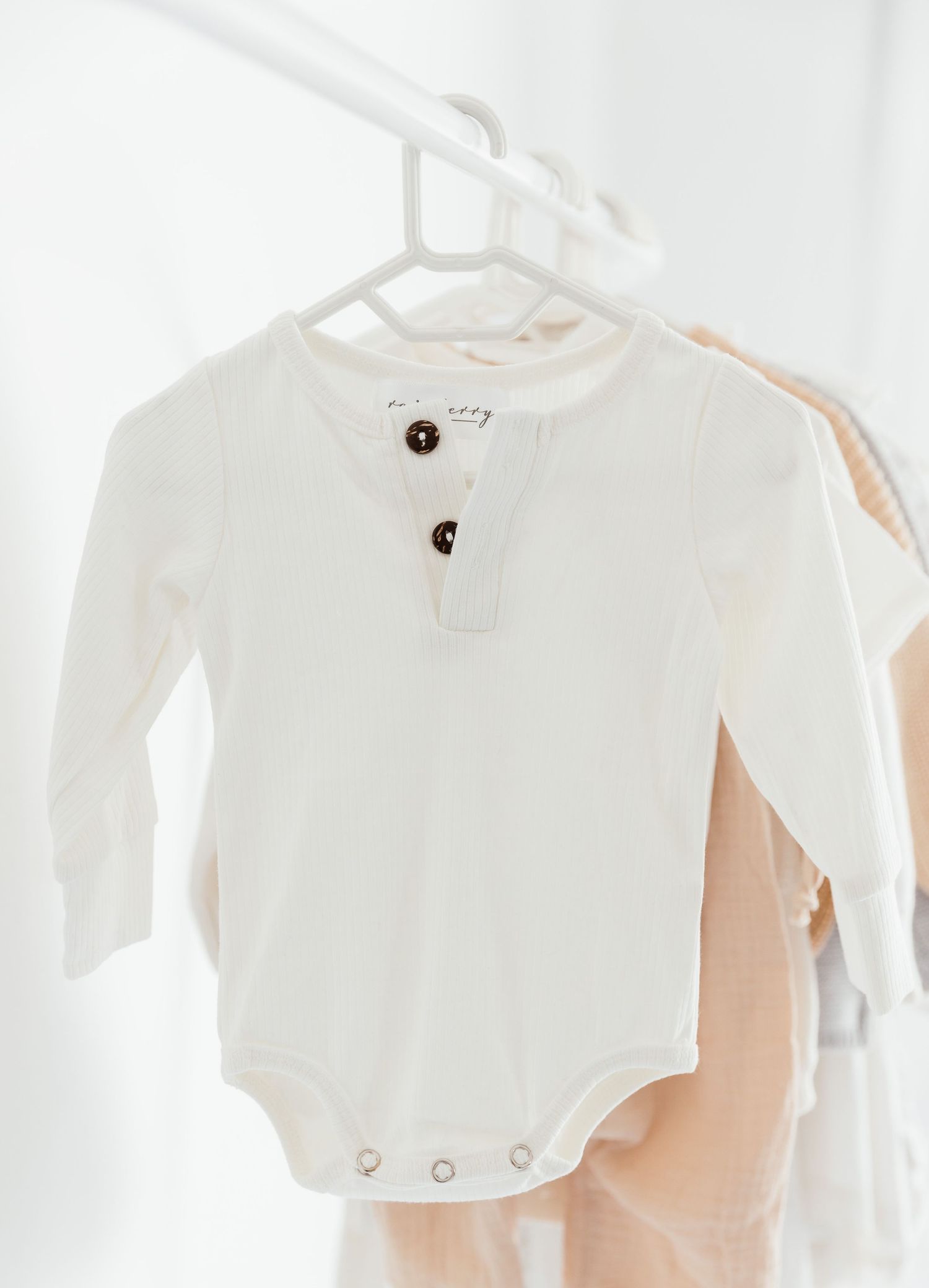 White long-sleeve baby bodysuit with brown buttons hanging on a hanger against a bright white background.