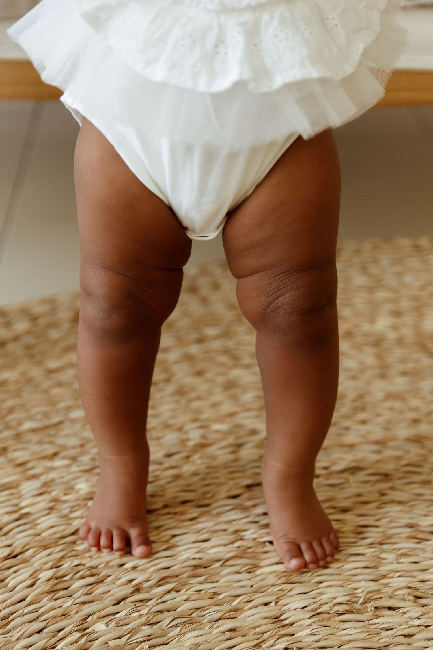 Close-up of chubby baby legs standing barefoot on a woven rug, wearing a white frilly dress and diaper.