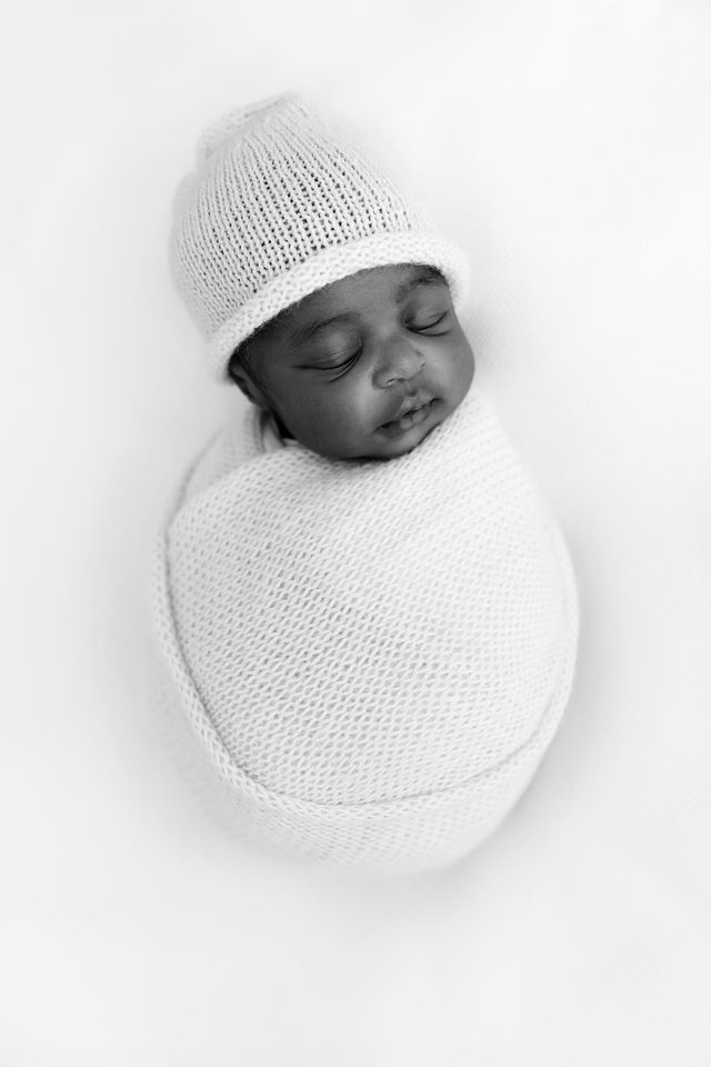 Black and white photo of a sleeping newborn baby swaddled in a knit wrap and wearing a small knit hat.