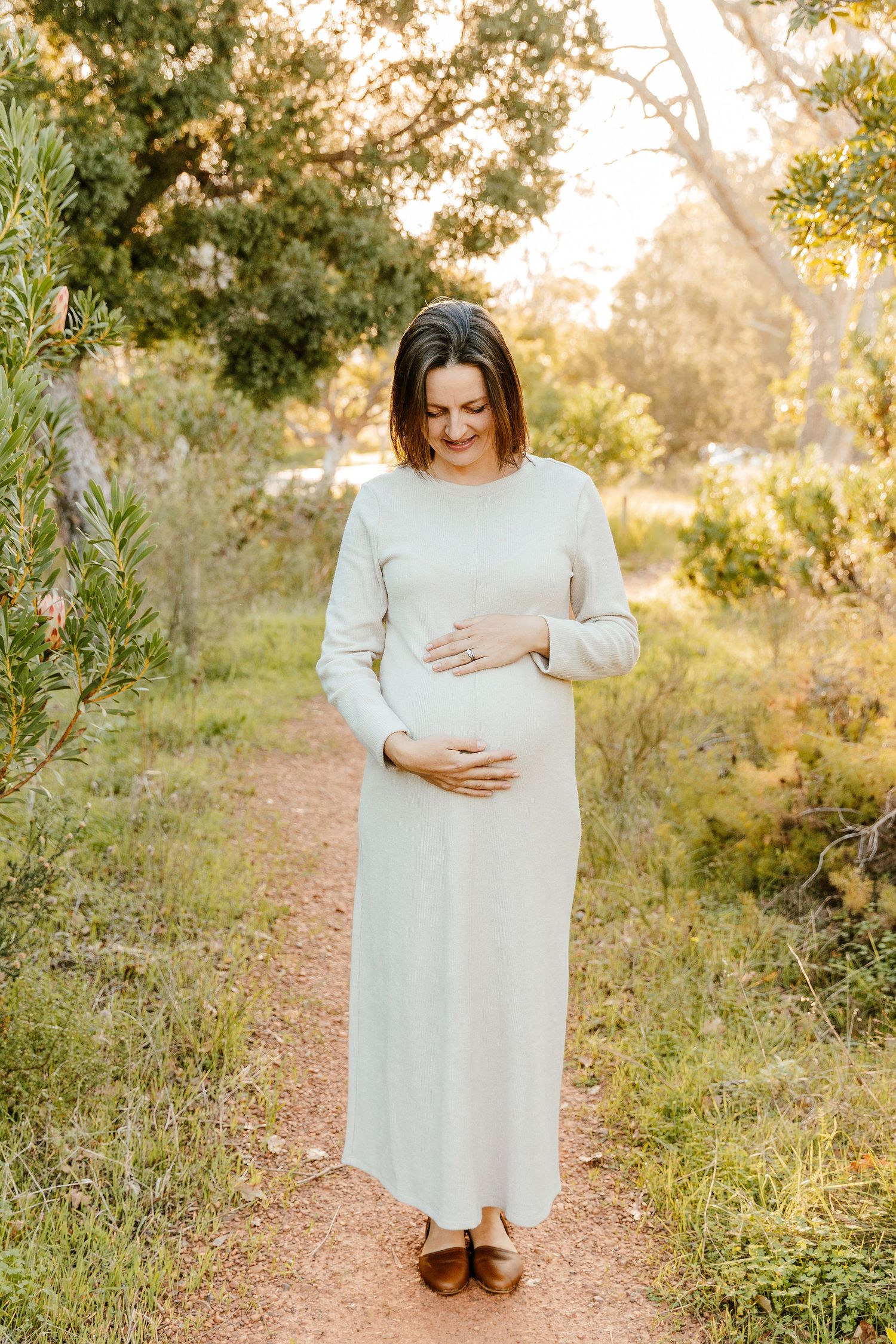 Pregnant woman in white long-sleeve dress cradling baby bump on nature path surrounded by green trees.