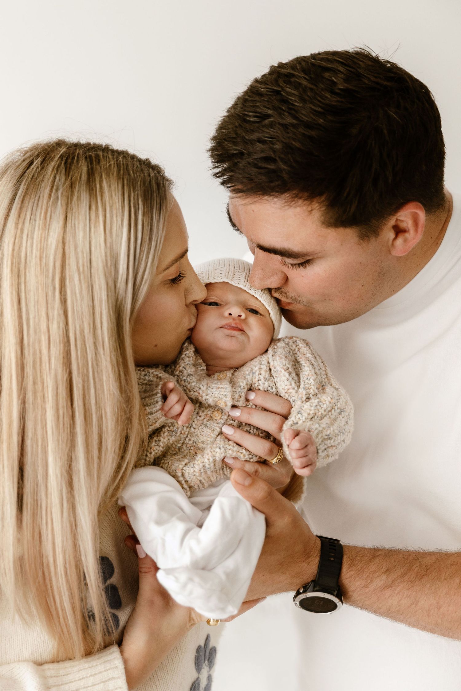 Both parents kissing their newborn baby on the cheeks simultaneously.