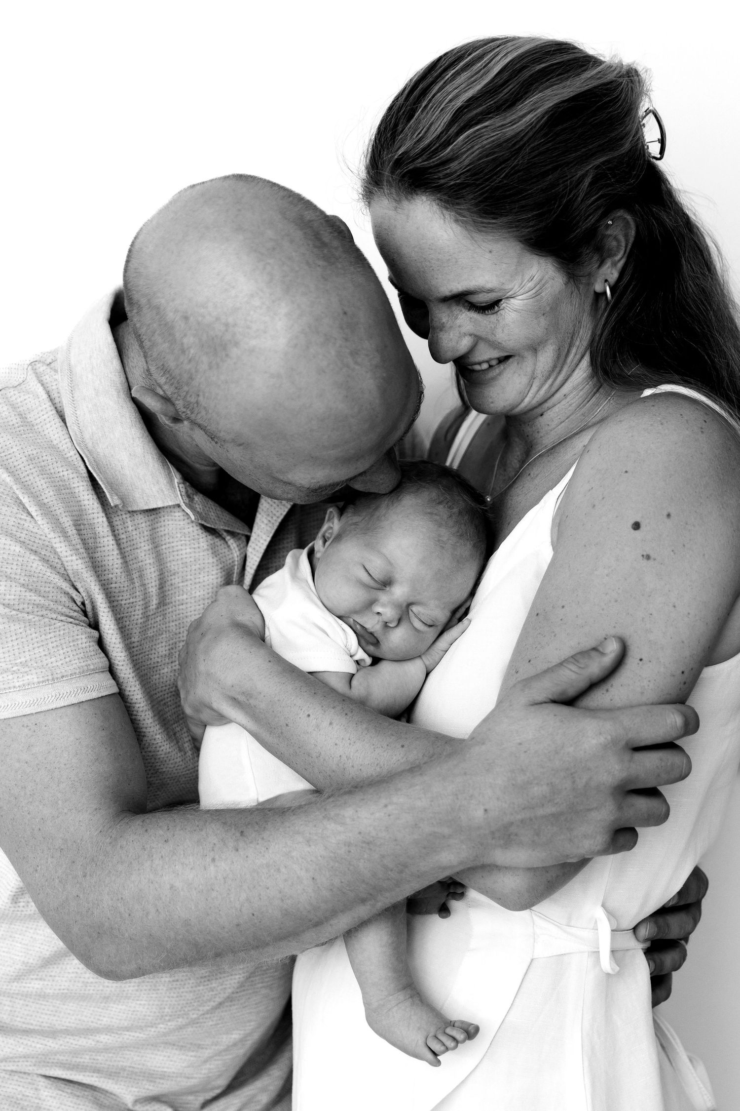 Black and white photo of parents lovingly cradling their sleeping newborn baby together in a tender family portrait.