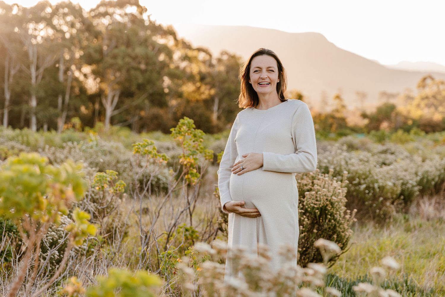 Smiling pregnant woman in white dress cradling baby bump in golden hour field with mountain backdrop.