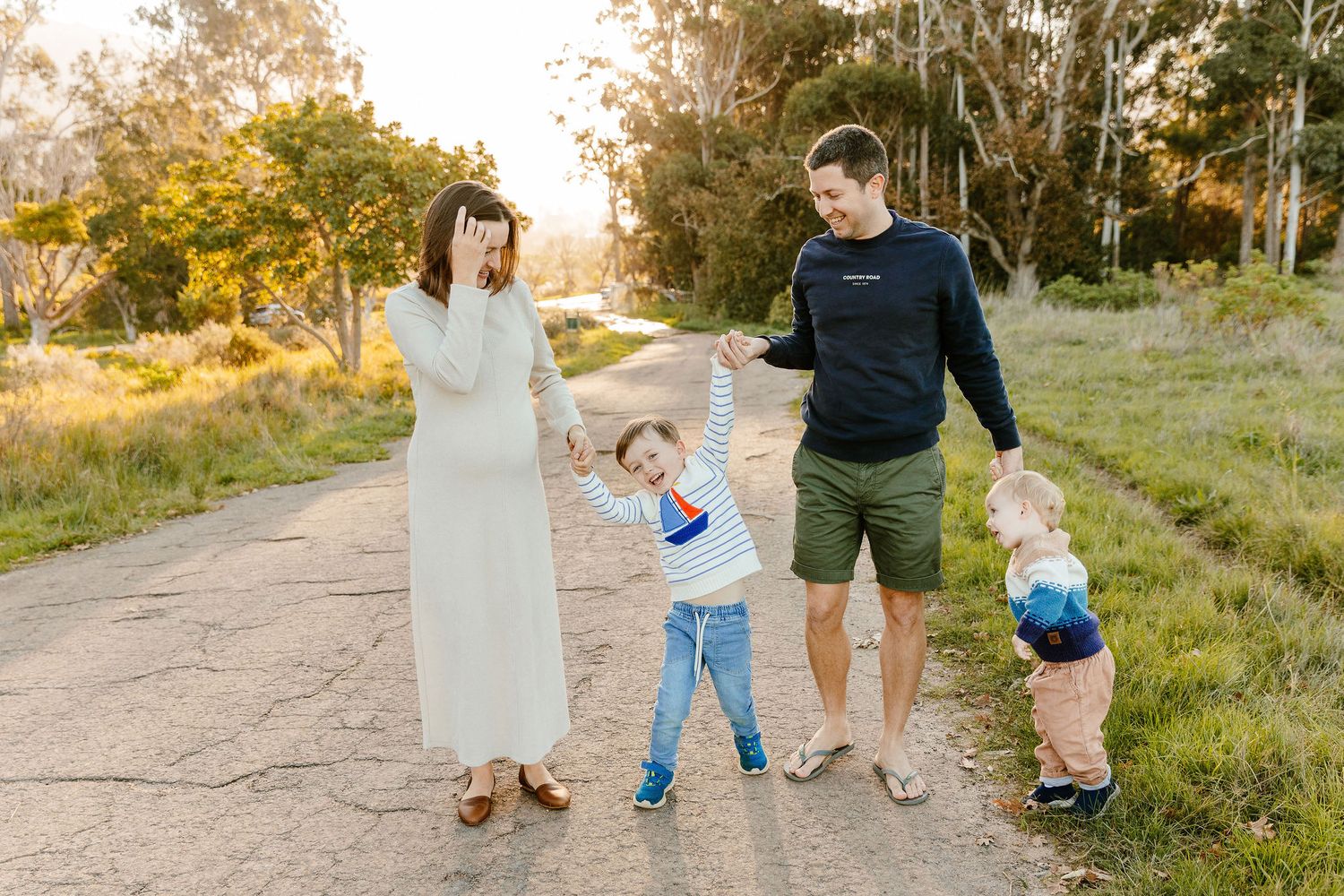 Joyful family of four playing on a sunny path, pregnant mom covering face while dad swings child outdoors.