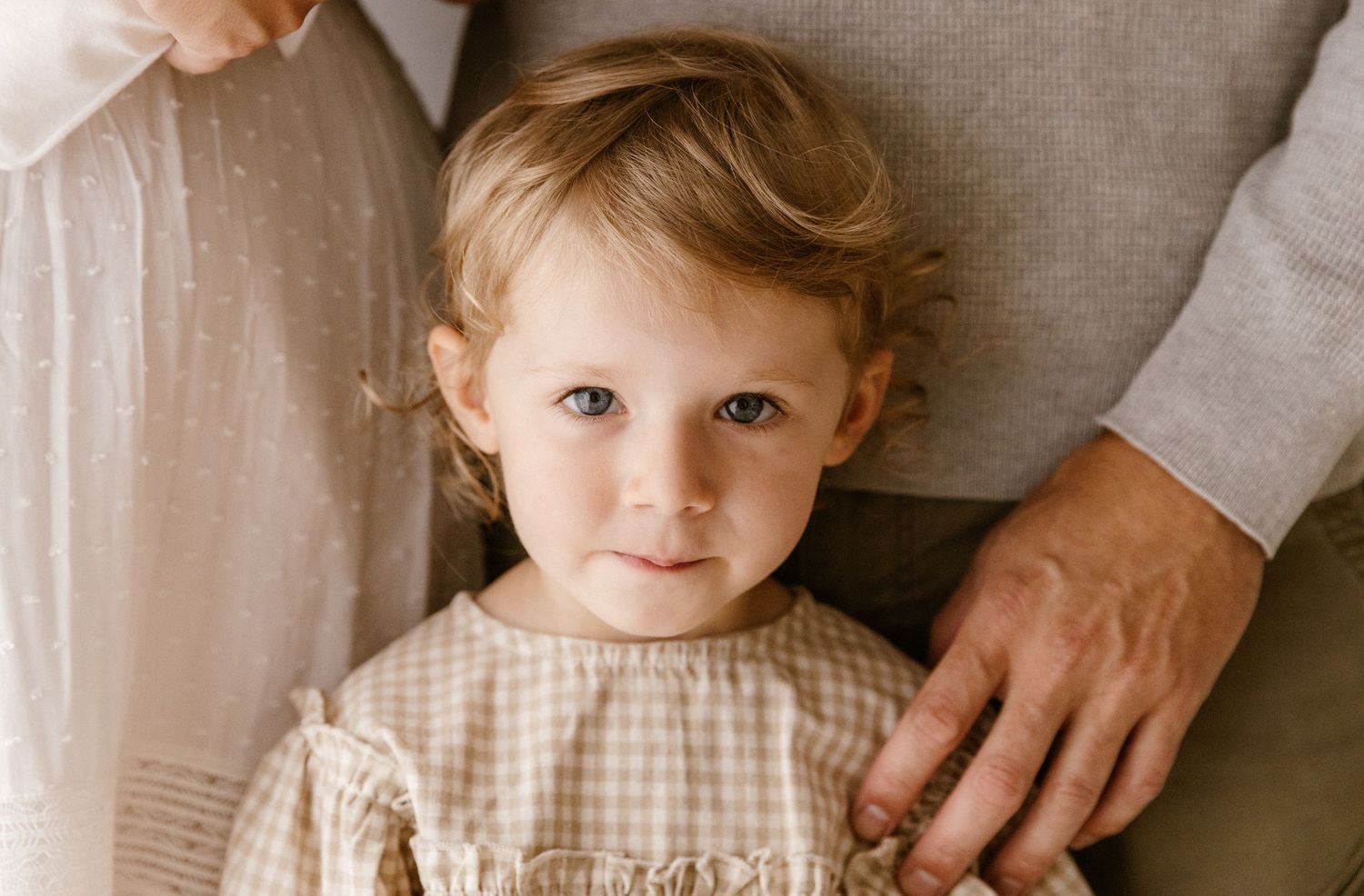 A young toddler with curly blonde hair and blue eyes in a gingham dress stands between two adults.