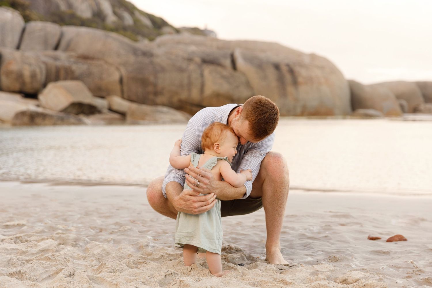 Father crouching on sandy beach kissing baby while gentle waves and rocky boulders create a serene backdrop.