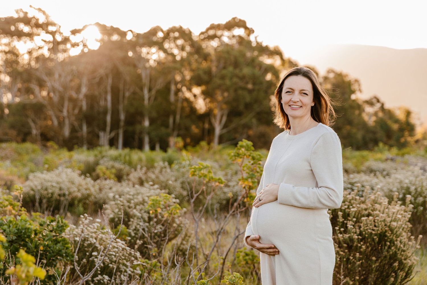 Smiling pregnant woman in white dress cradling baby bump in golden hour sunlit meadow with eucalyptus trees.