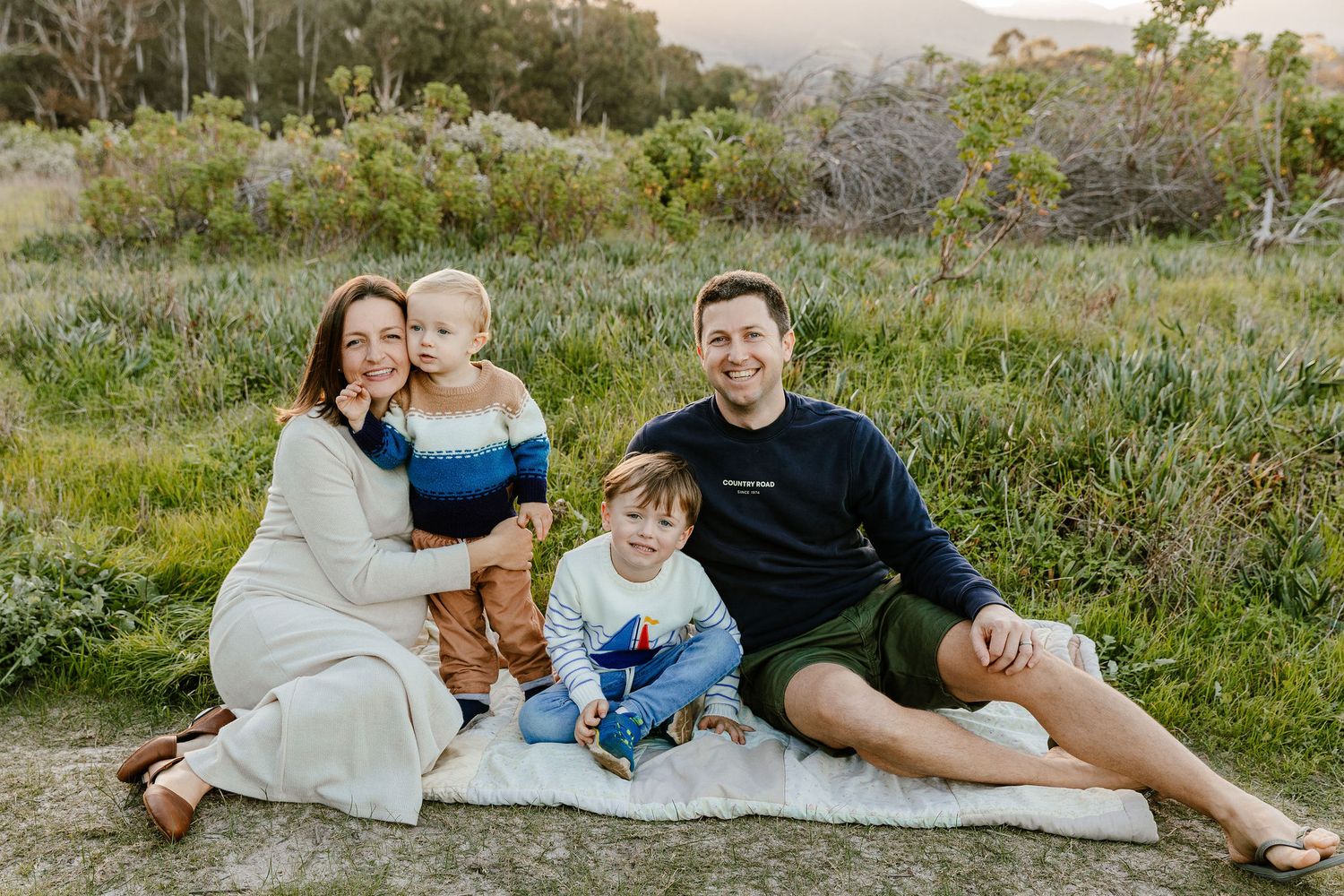 Smiling family of four sitting on a white blanket in a lush green outdoor setting.