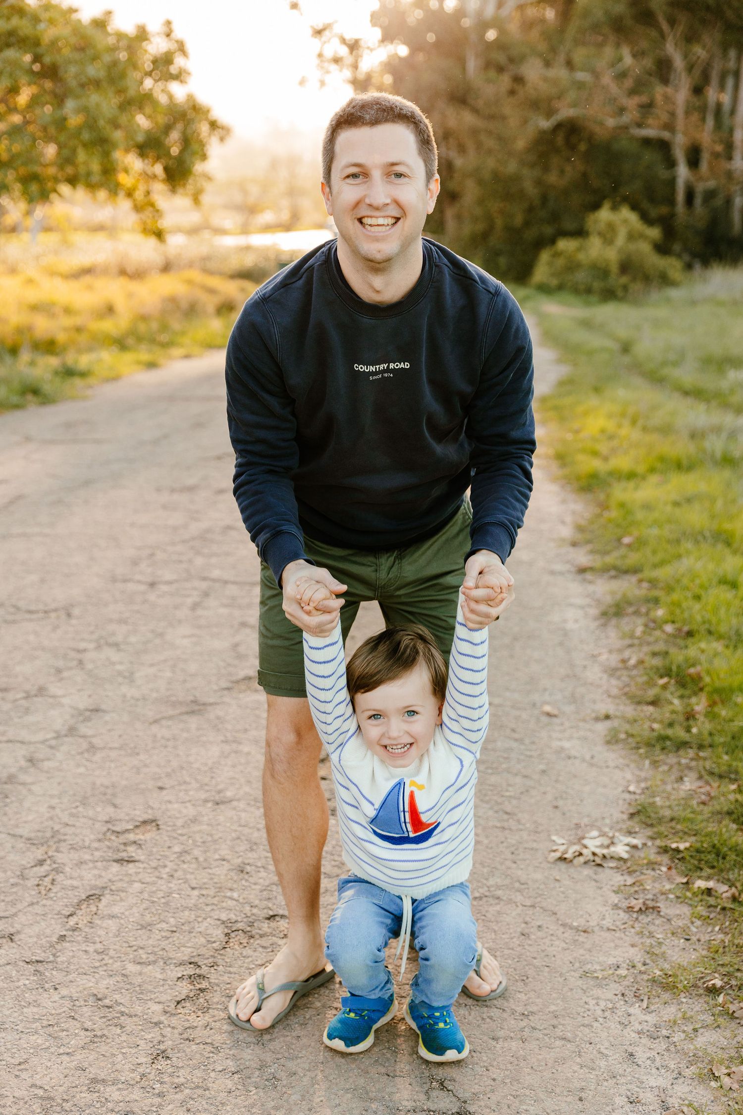 A happy father and his smiling toddler son pose together on a sunny outdoor dirt path.