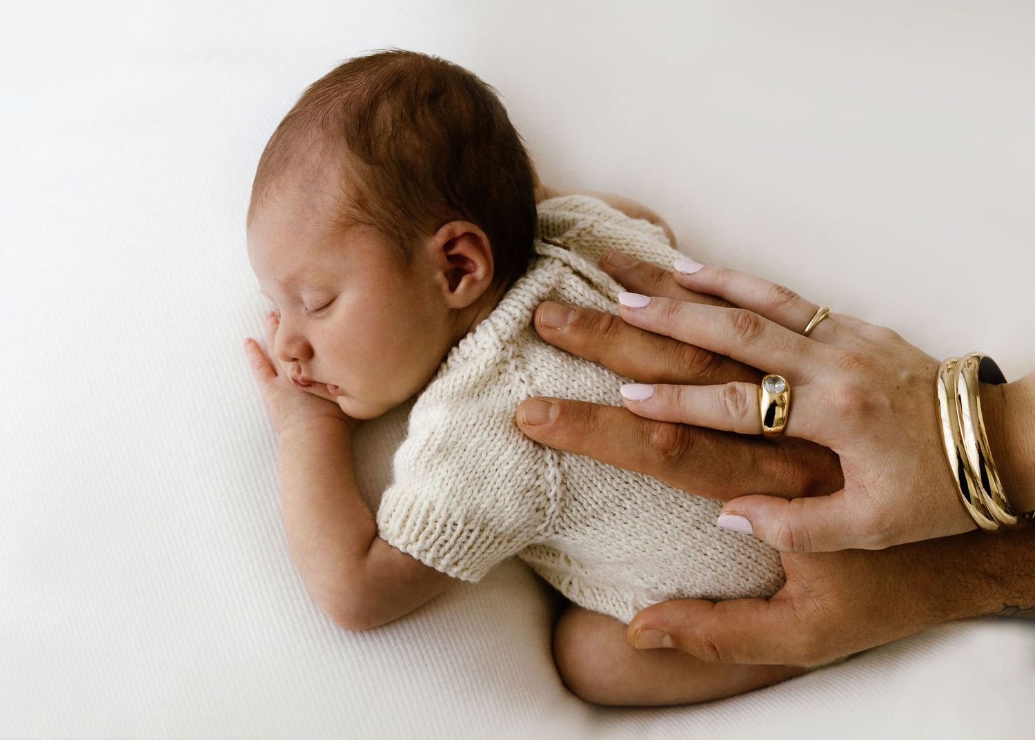 Parent's hands with gold rings tenderly holding sleeping newborn baby in cream knit outfit.