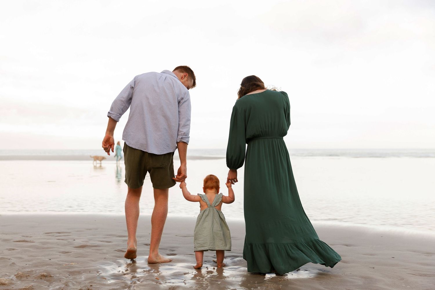 Family of three holding hands on a beach, parents and toddler girl in green dress facing the ocean.