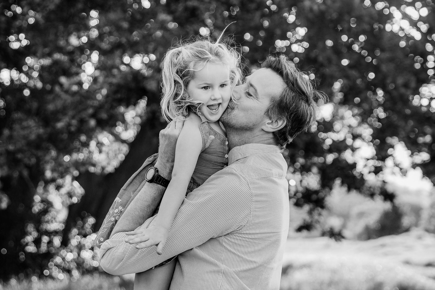 Black and white photo of a father kissing his young daughter on the cheek outdoors.