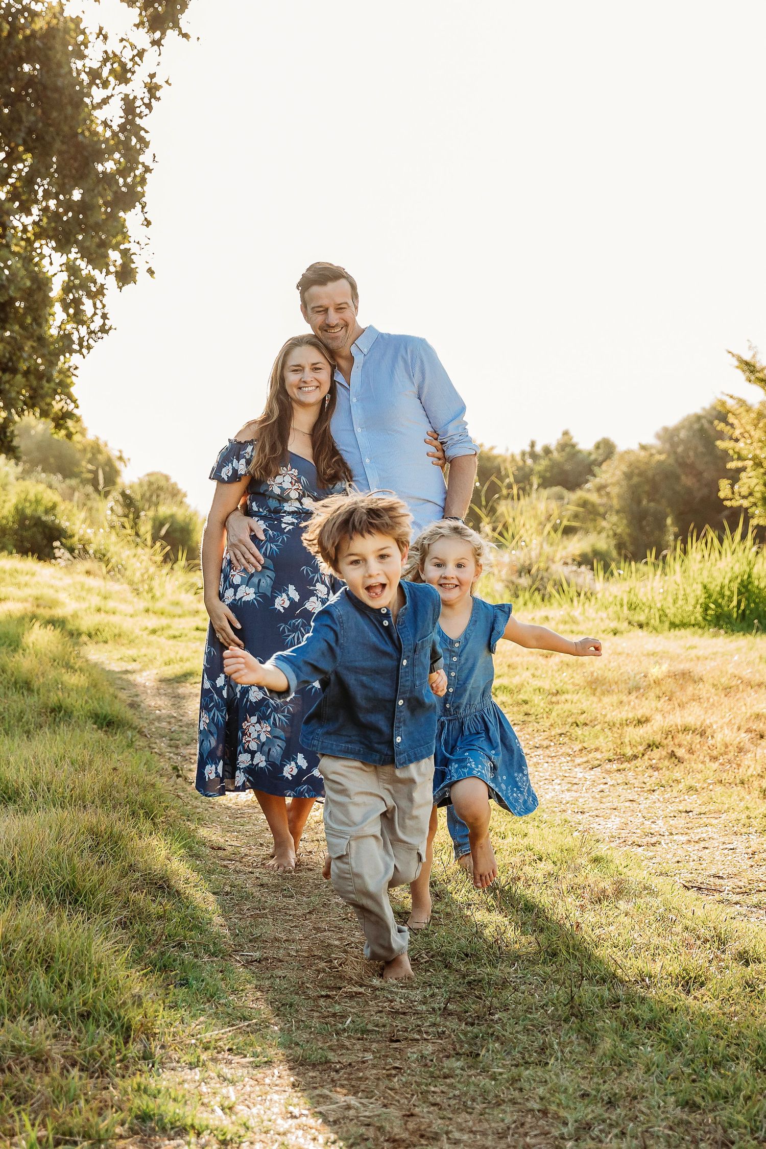 Happy family of four walking on a sunny path, parents in blue outfits with two young children running ahead.
