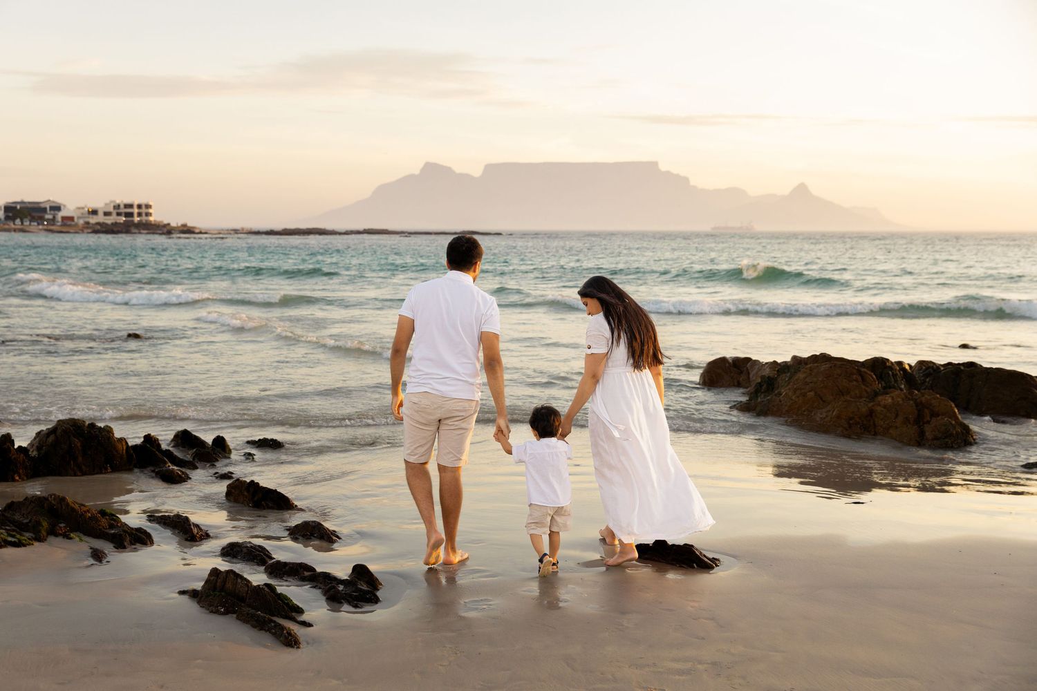 Family of three holding hands on a sandy beach at sunset with Table Mountain in the background.
