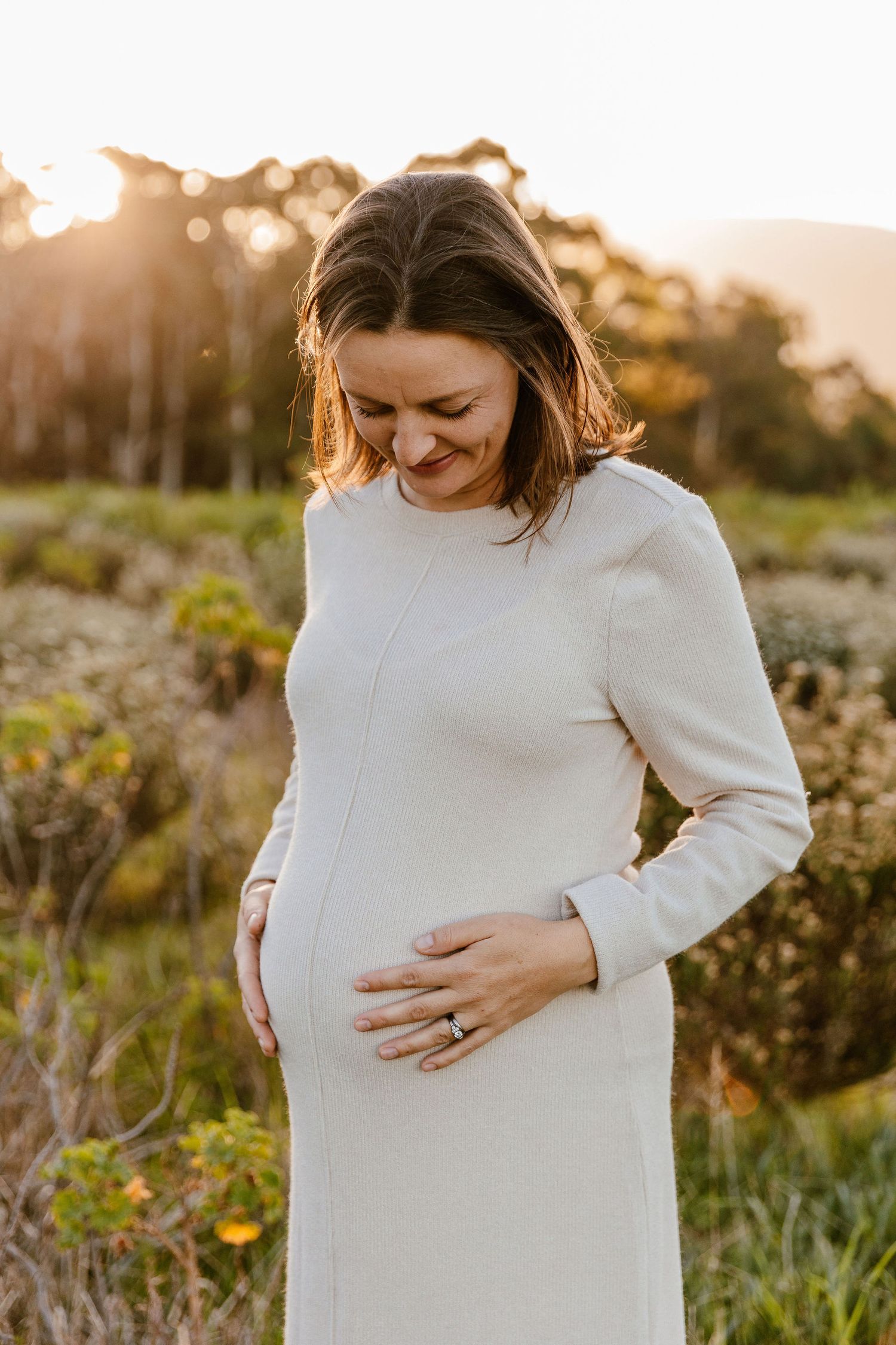 Pregnant woman in white dress cradling baby bump outdoors at golden hour sunset.