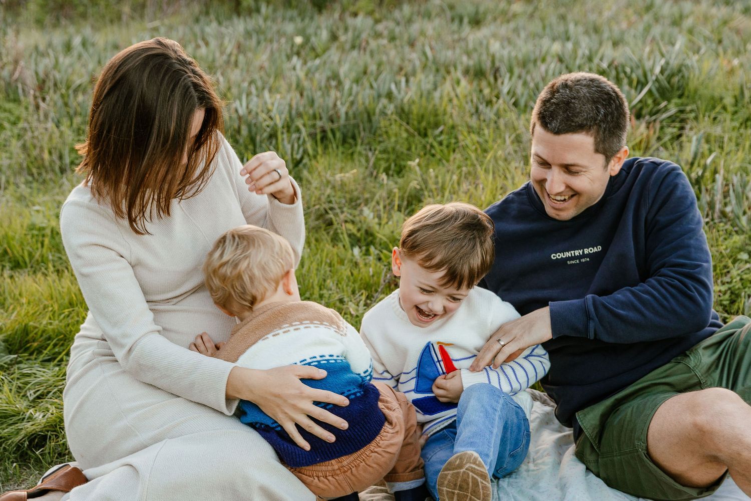 Happy family of four enjoying a picnic on a blanket in a lush green field, laughing and playing together.