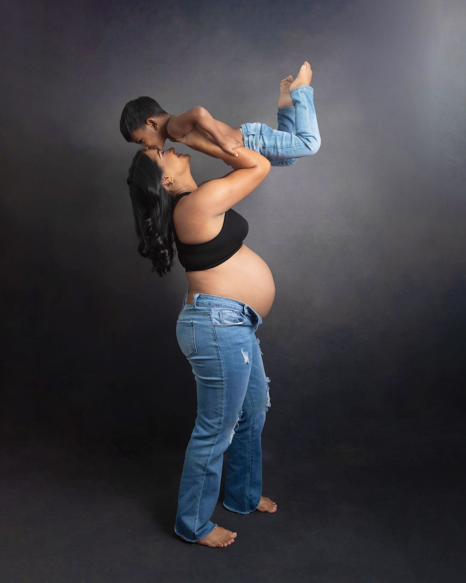 Pregnant mom in casual wear lifting her toddler and photographed against a charcoal backdrop