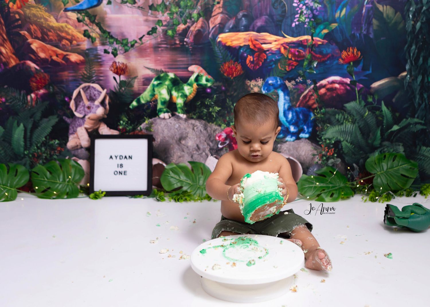 Dinosaur themed cakesmash with baby lifting cake up off the stand