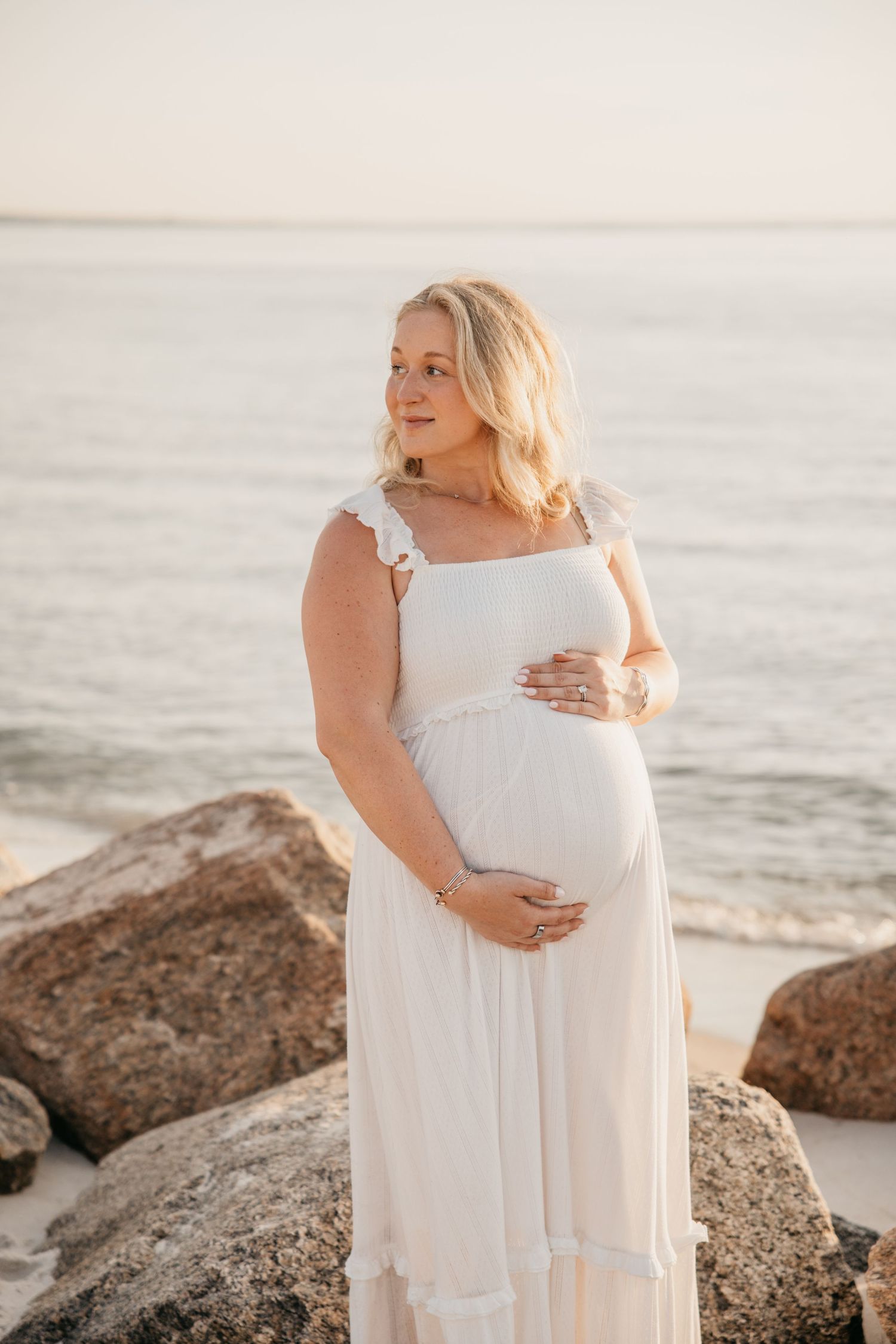 Pregnant woman in white dress cradling baby bump on rocky beach at golden hour sunset.