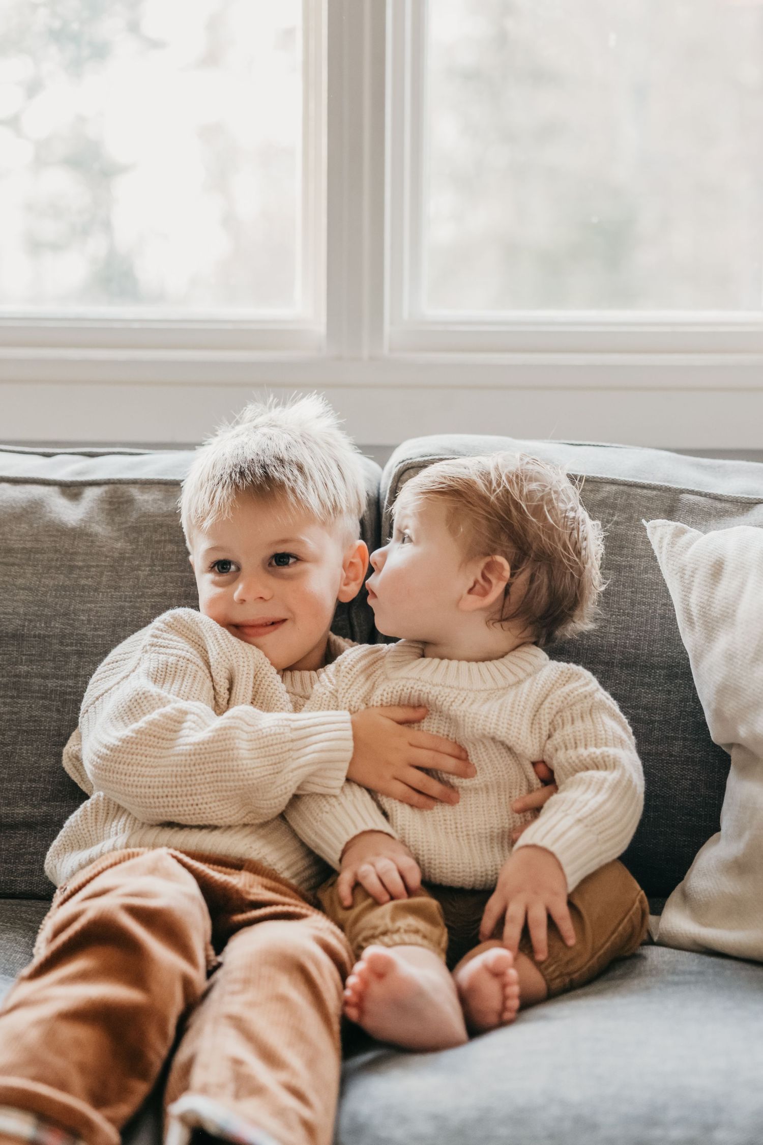 Two small children in matching sweaters share sweet moment on gray couch.
