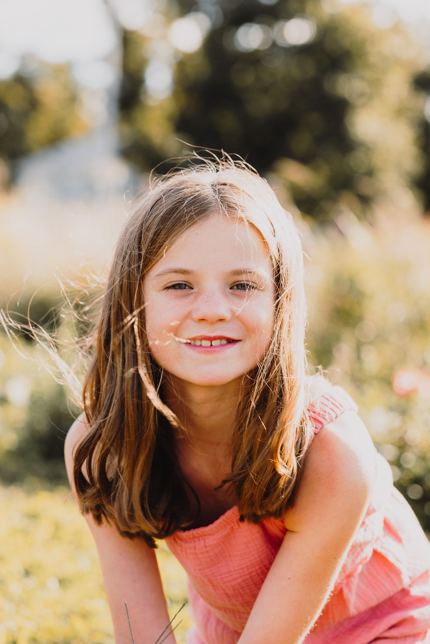 Child in pink dress smiles brightly in a sun-drenched summer garden.