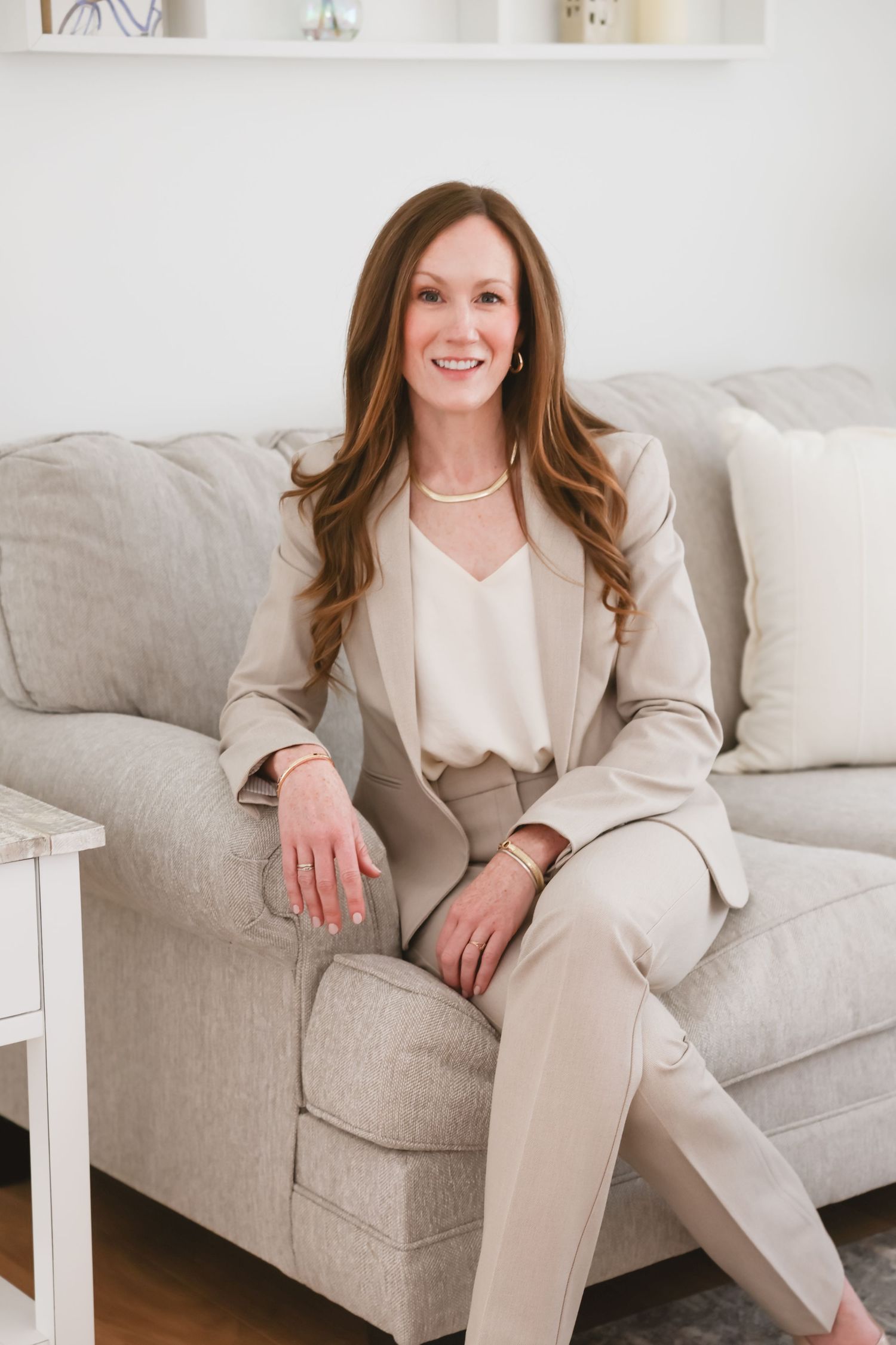 A smiling woman in a beige suit sits elegantly on a gray sofa in a bright, modern living room.