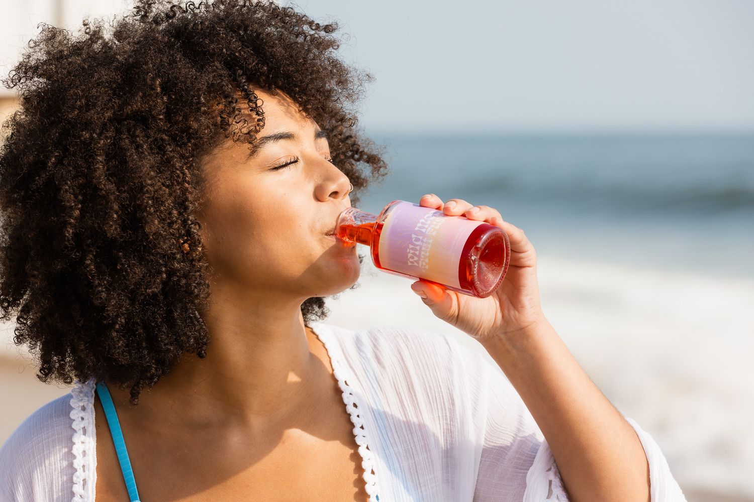 Person with curly hair enjoying a pink drink at the beach.