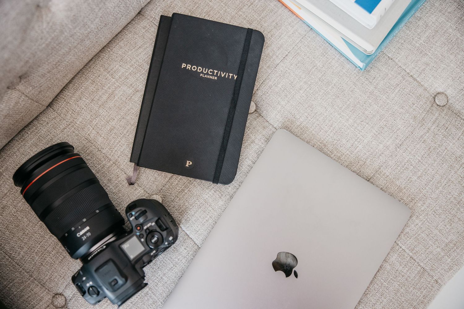 Camera, Productivity Planner notebook, and MacBook laptop arranged on a beige linen sofa for a creative workspace flatlay.