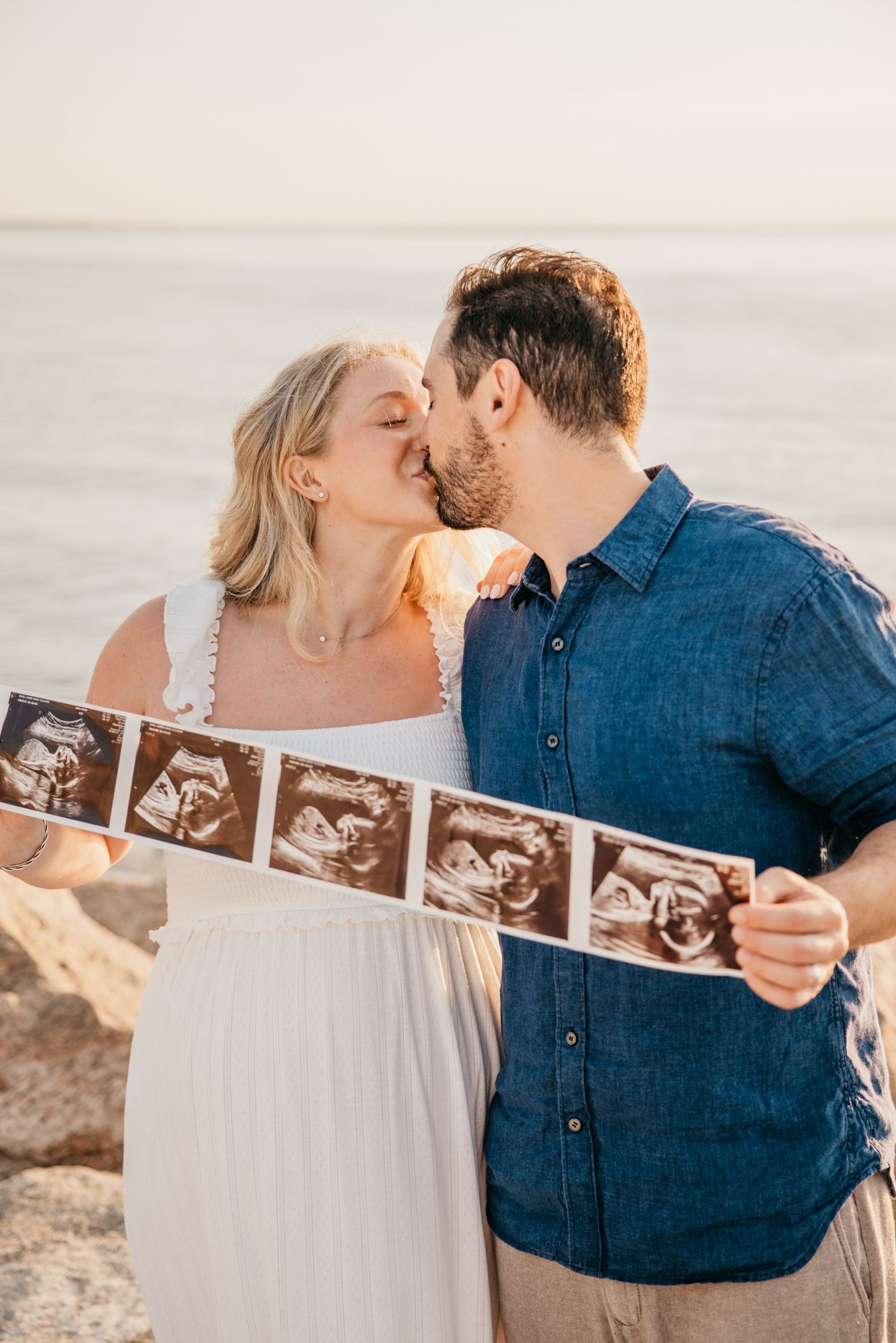 Couple kissing on beach holding ultrasound photos, pregnancy announcement with ocean backdrop at golden hour.