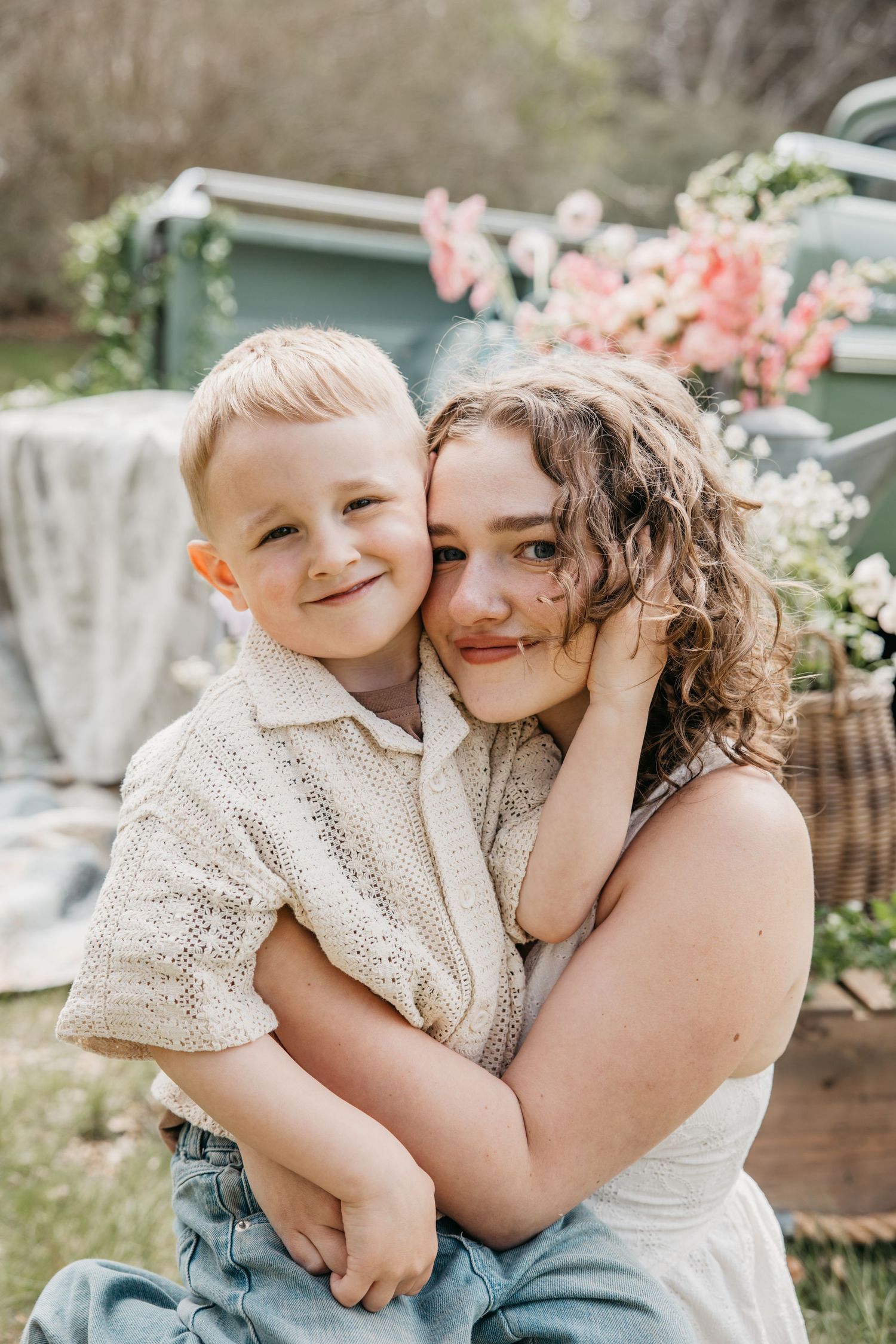 A young boy hugs a curly-haired woman outdoors near floral decorations in a warm, sunny garden setting.