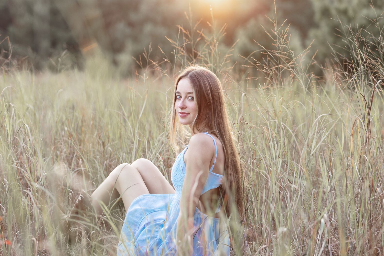 Person in light blue dress sits gracefully among tall grass at golden hour.