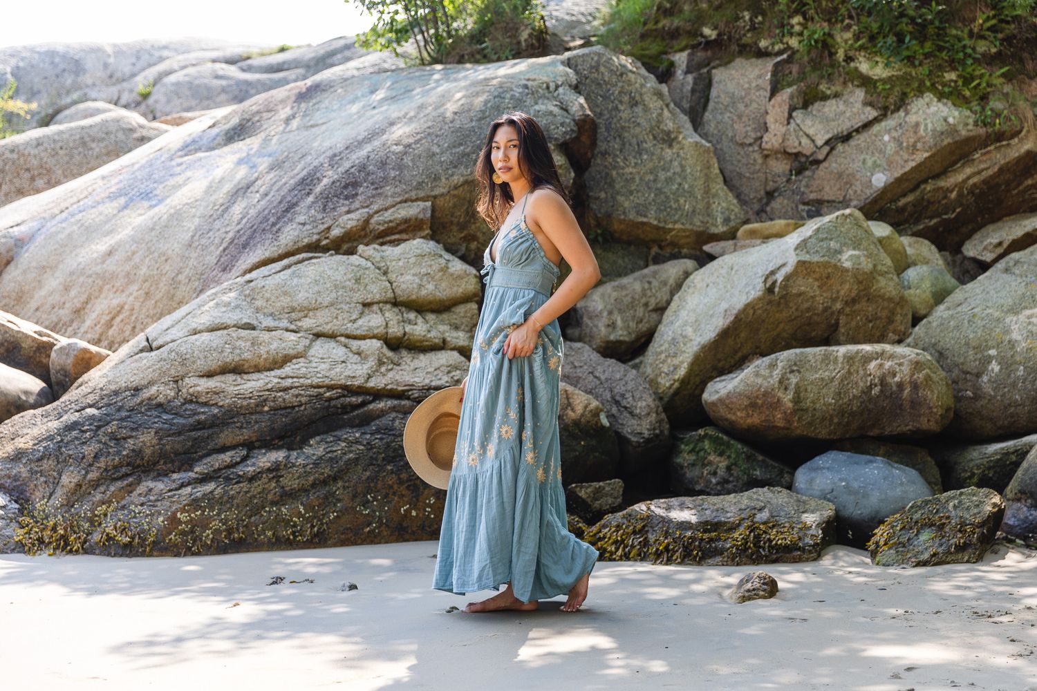 Woman in blue dress poses gracefully on rocky beach during outdoor photo session.