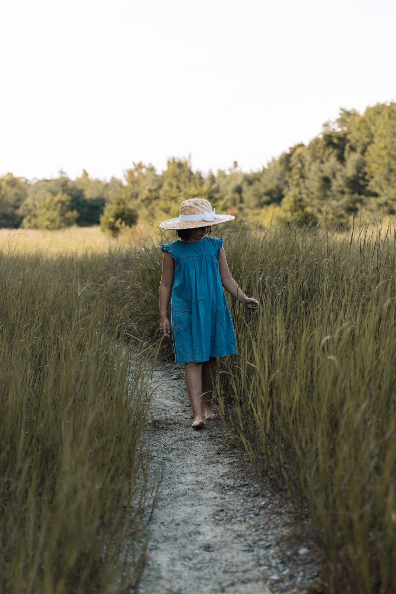 Person walking along a sandy path through tall grass at sunset.