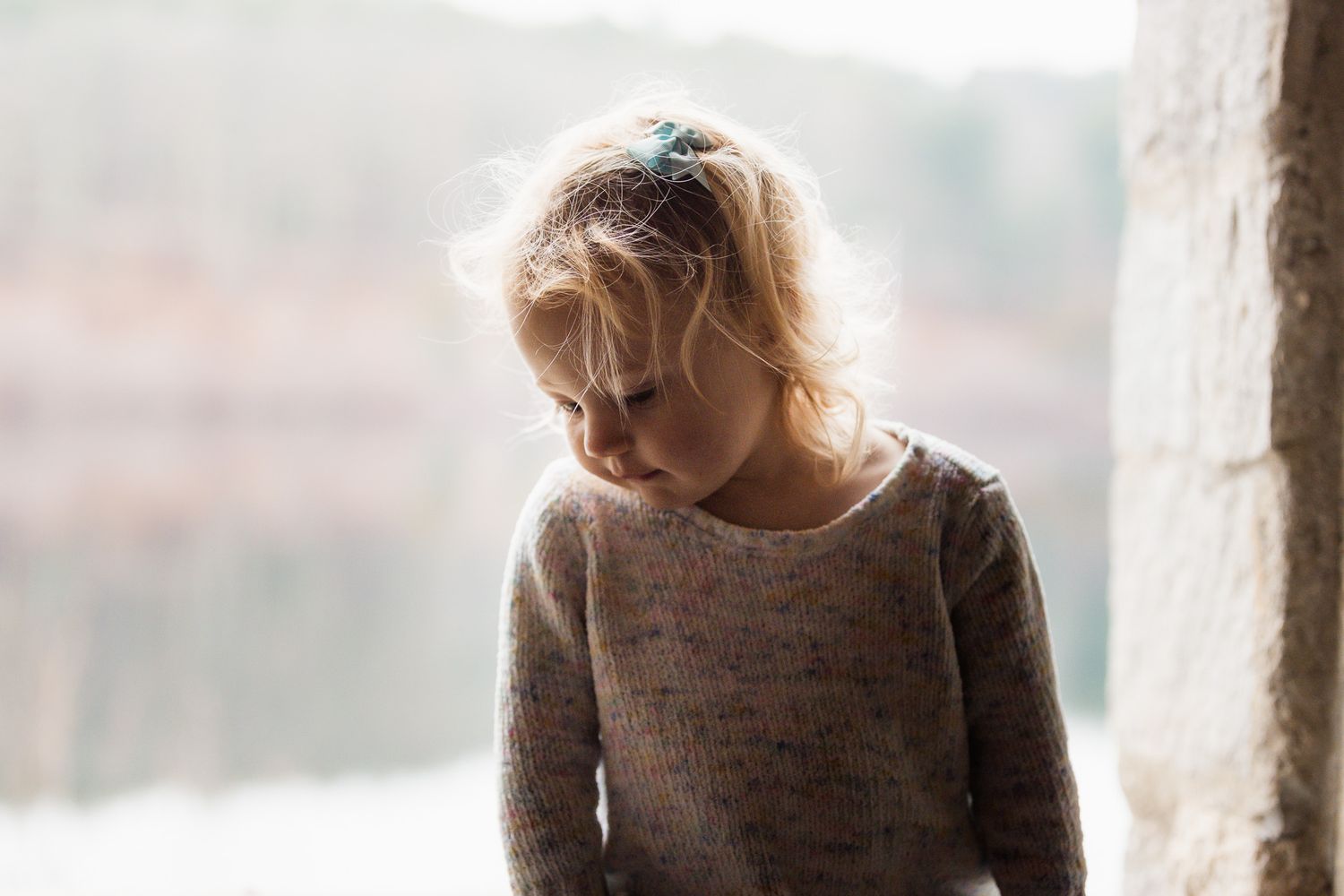 Person in brown sweater looking down thoughtfully outdoors.