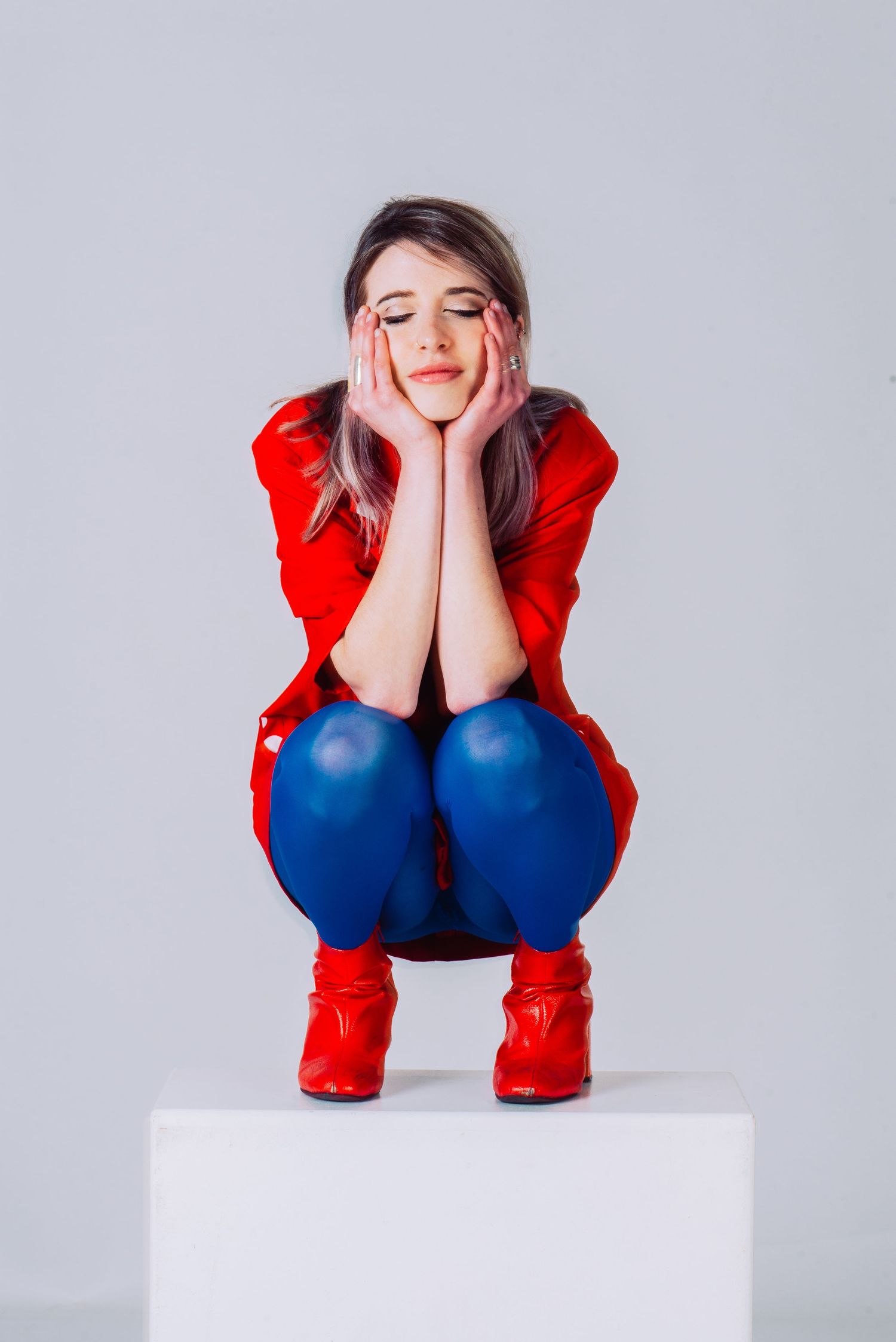 Person wearing red top and blue pants crouching on white platform in studio setting.