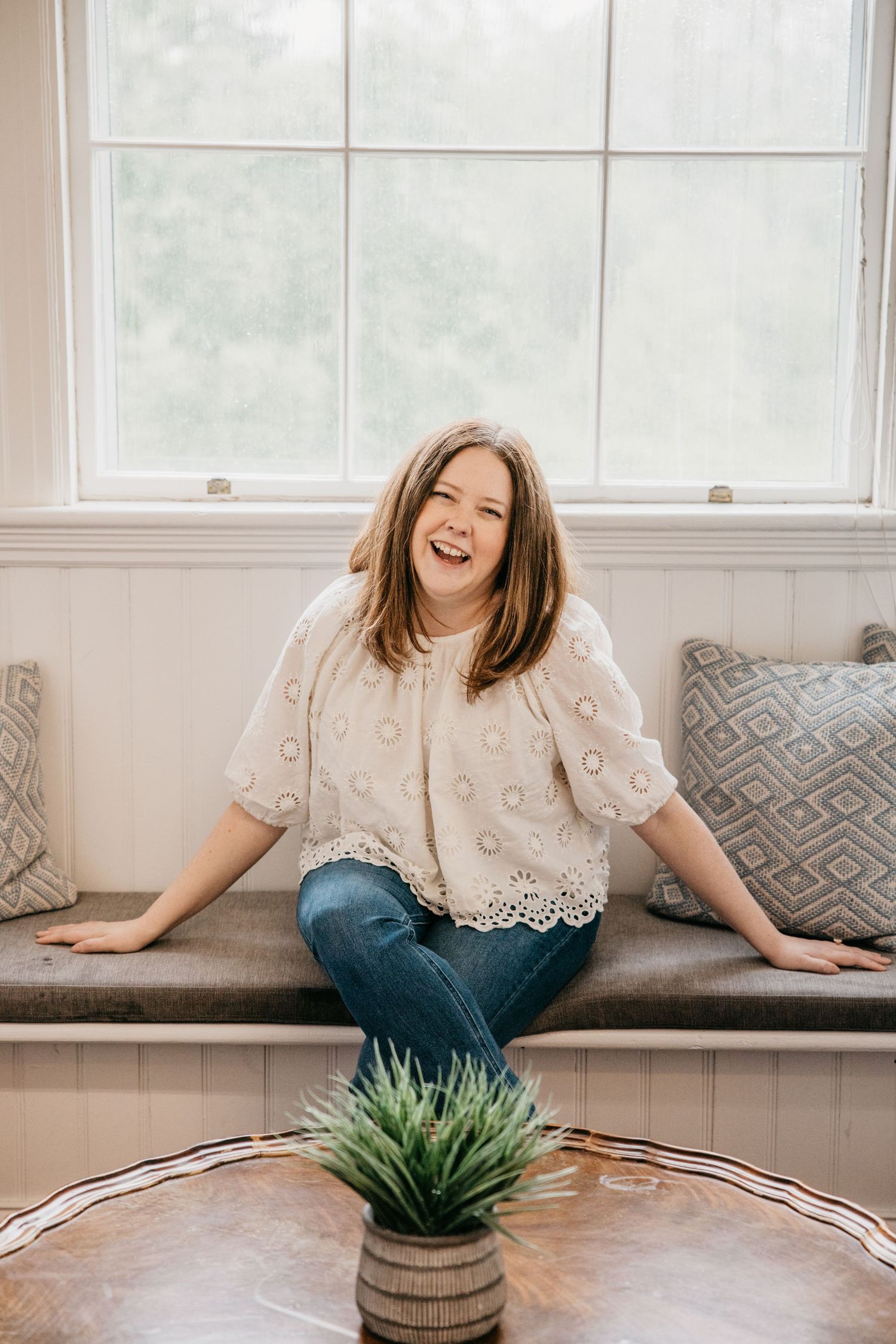 A smiling woman sits cross-legged on a window bench, wearing a white lace top and jeans, with a small plant in the foreground.