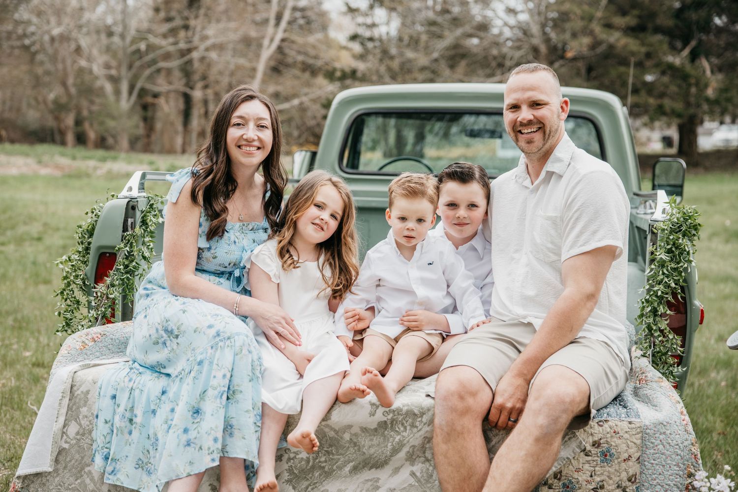 Happy family of five sitting on vintage green truck tailgate outdoors, smiling in spring setting.