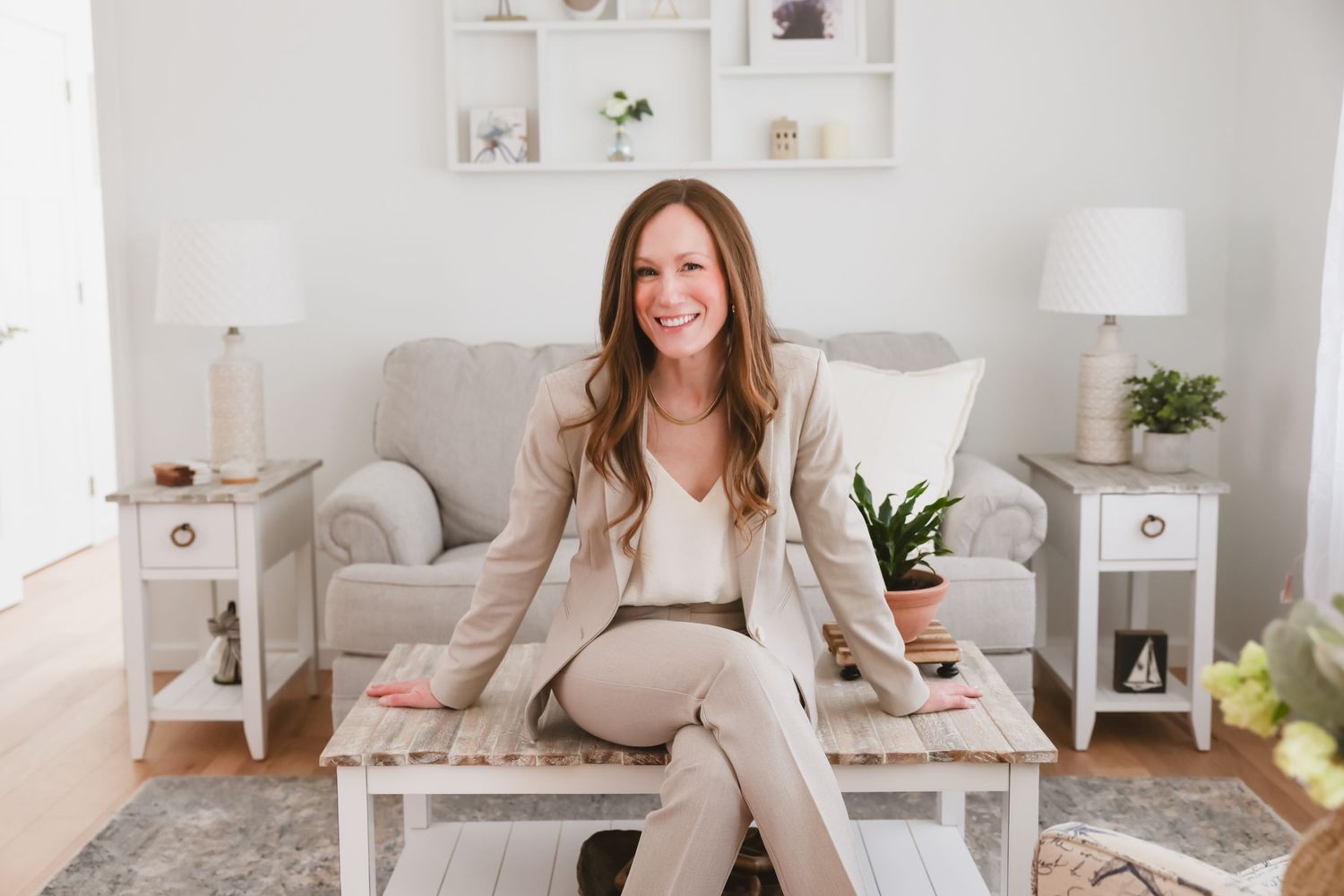 Smiling woman in beige blazer sitting on white coffee table in bright, modern living room with neutral decor.