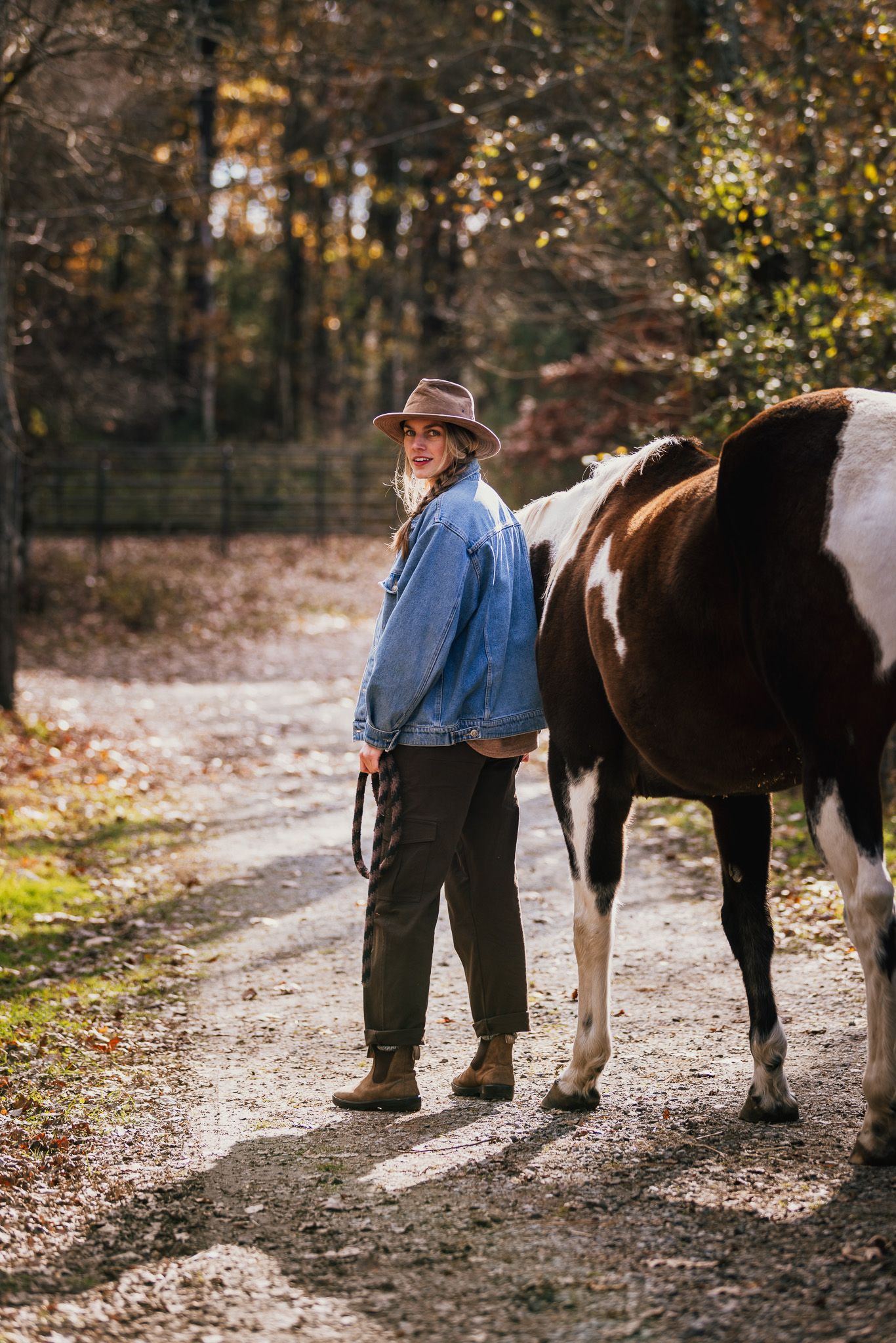 Person in casual attire standing next to horses on an autumn trail.