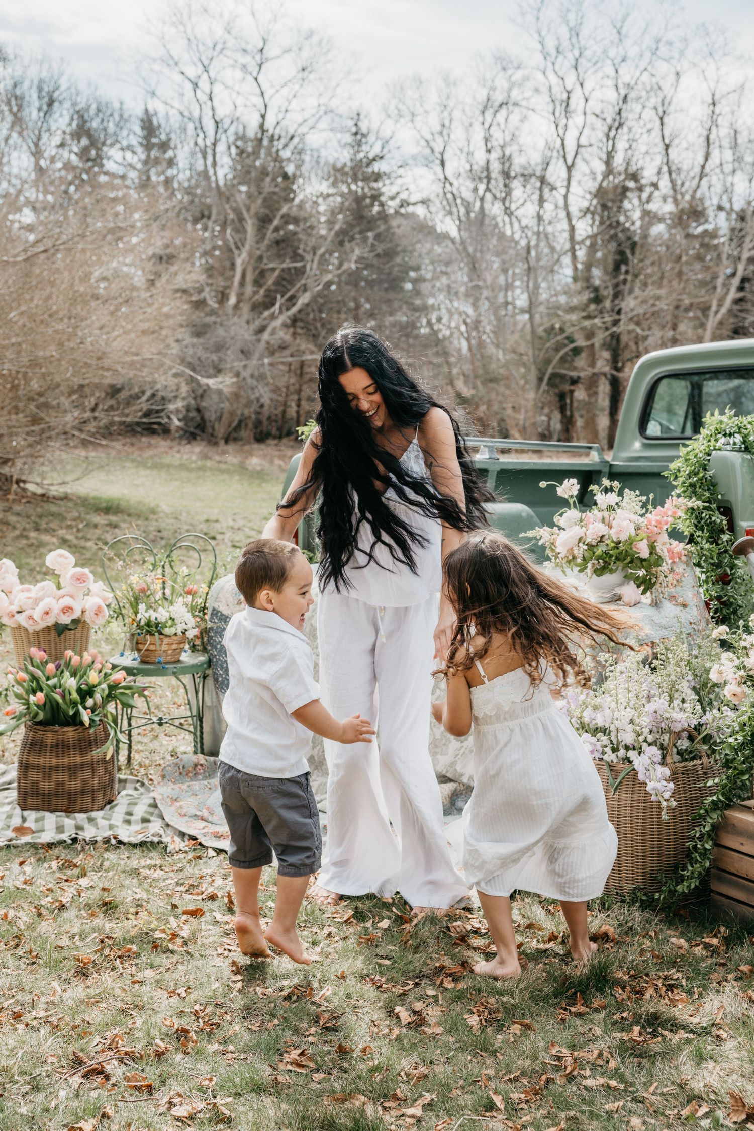 A woman in white dances outdoors with two children among floral arrangements near a vintage green truck.