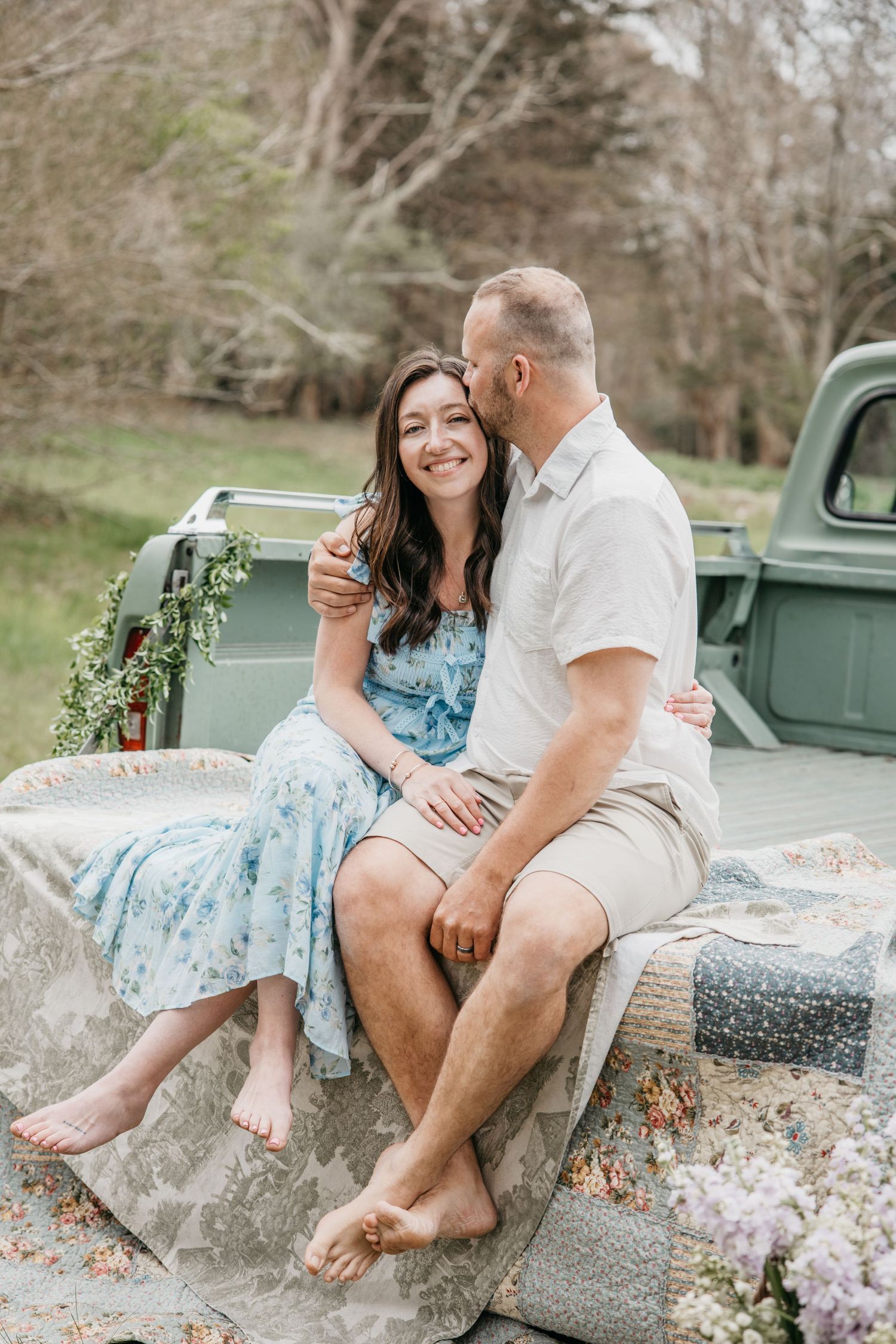 A couple sits barefoot on a vintage green truck bed, sharing a tender moment outdoors with a floral wreath in the background.