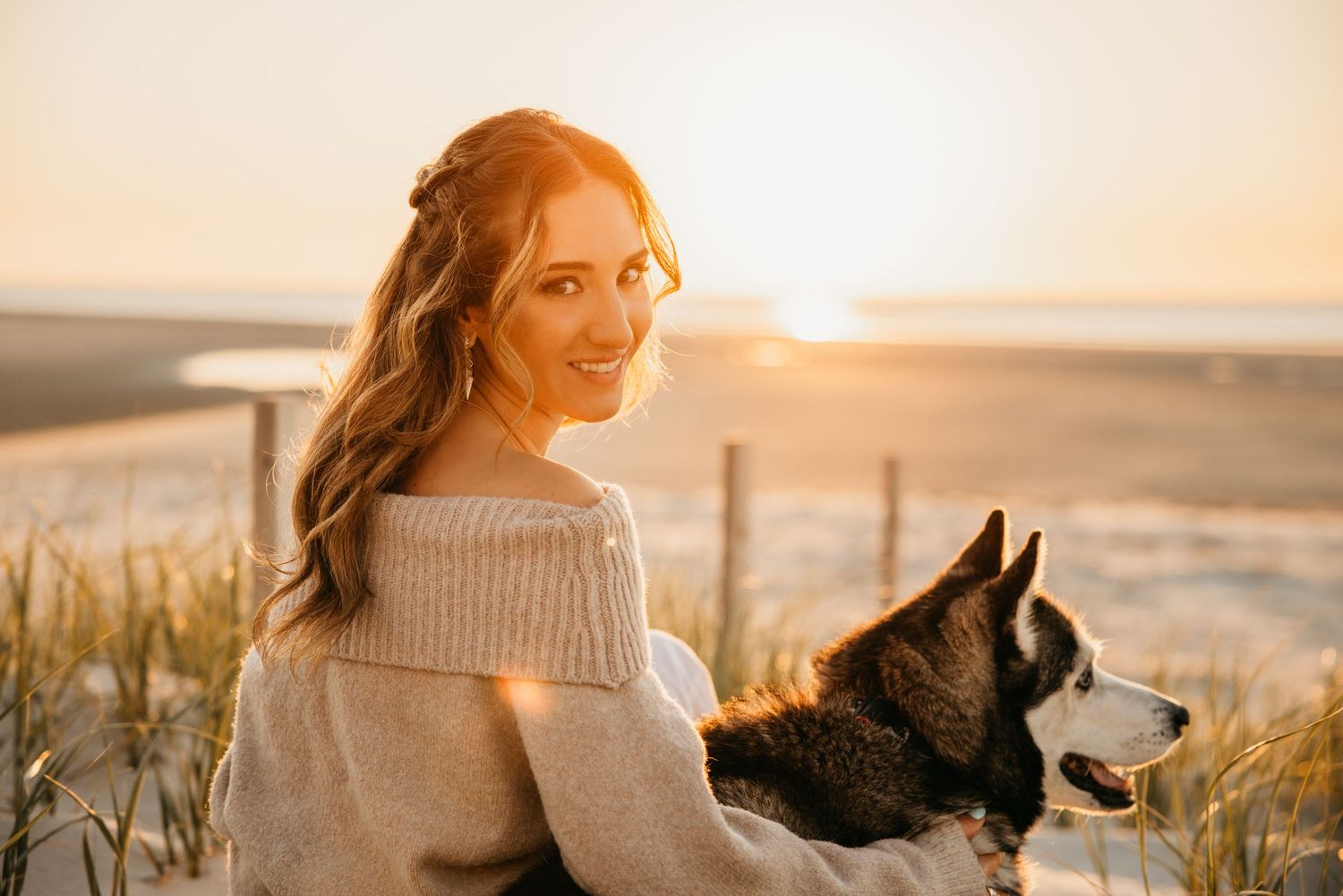 Woman in cozy sweater smiling at beach sunset with Husky dog, golden hour light glowing behind them.