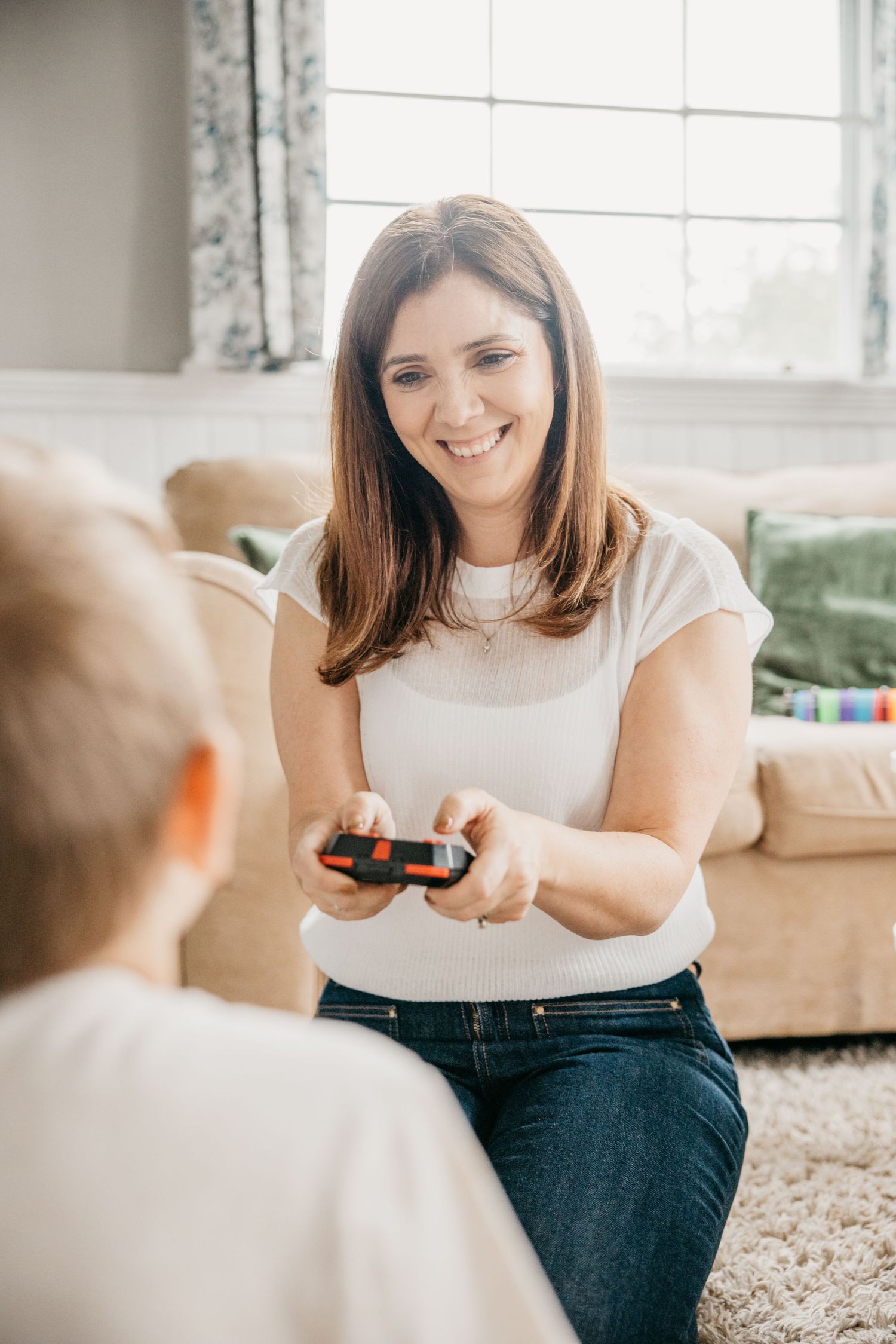 A smiling woman in a white top and jeans plays a video game with a child in a bright living room.