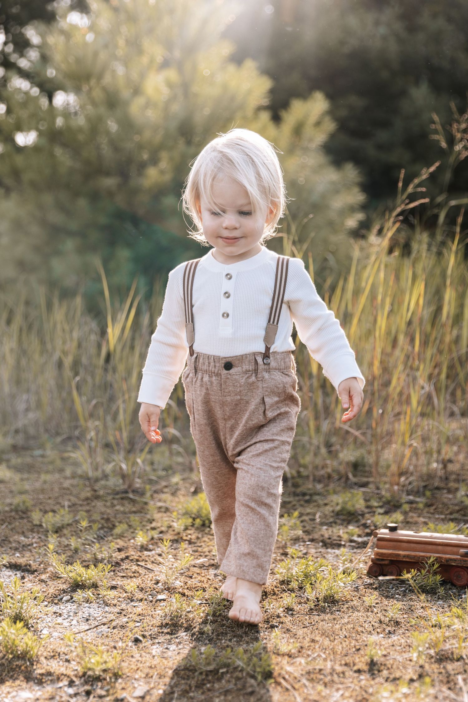 A toddler in brown overalls and white shirt walks through a sunny outdoor setting.