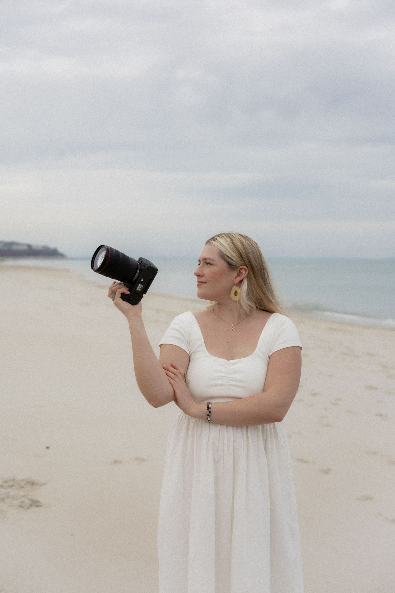 A photographer in a white dress stands on a sandy beach taking photos with a professional camera.